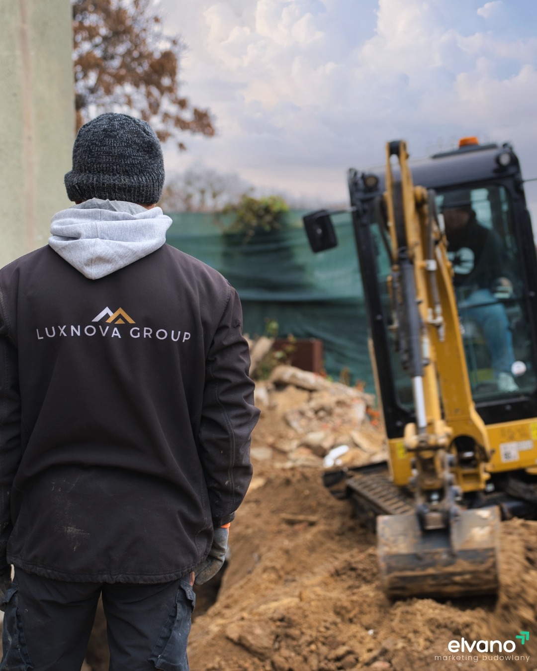 Mężczyzna w czarnej kurtce z logo Luxnova Group obserwuje pracę żółtej koparki na placu budowy, widoczne gruzy i wykopana ziemia.
