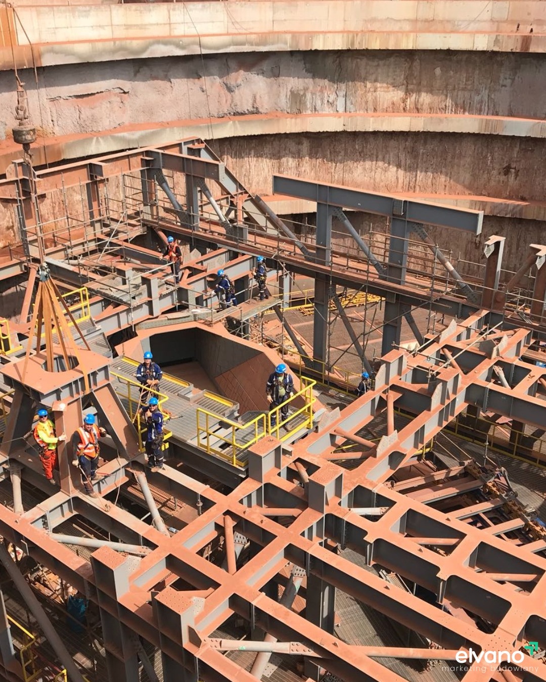 Konstrukcja stalowa w głębokim wykopie, pracownicy w kaskach i odzieży ochronnej na platformach roboczych, widok z góry na szkielet budowli.