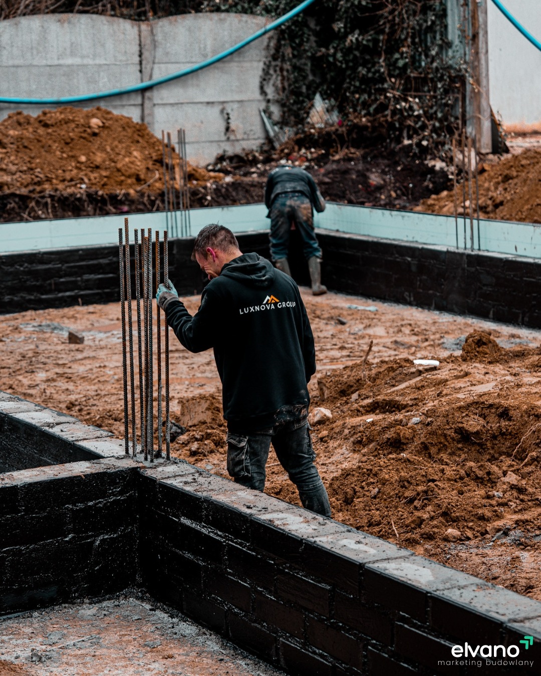 Budowa fundamentów domu: robotnicy w trakcie wylewania betonu i montażu zbrojenia, widoczny fragment ściany pokrytej czarnym hydroizolacyjnym preparatem.