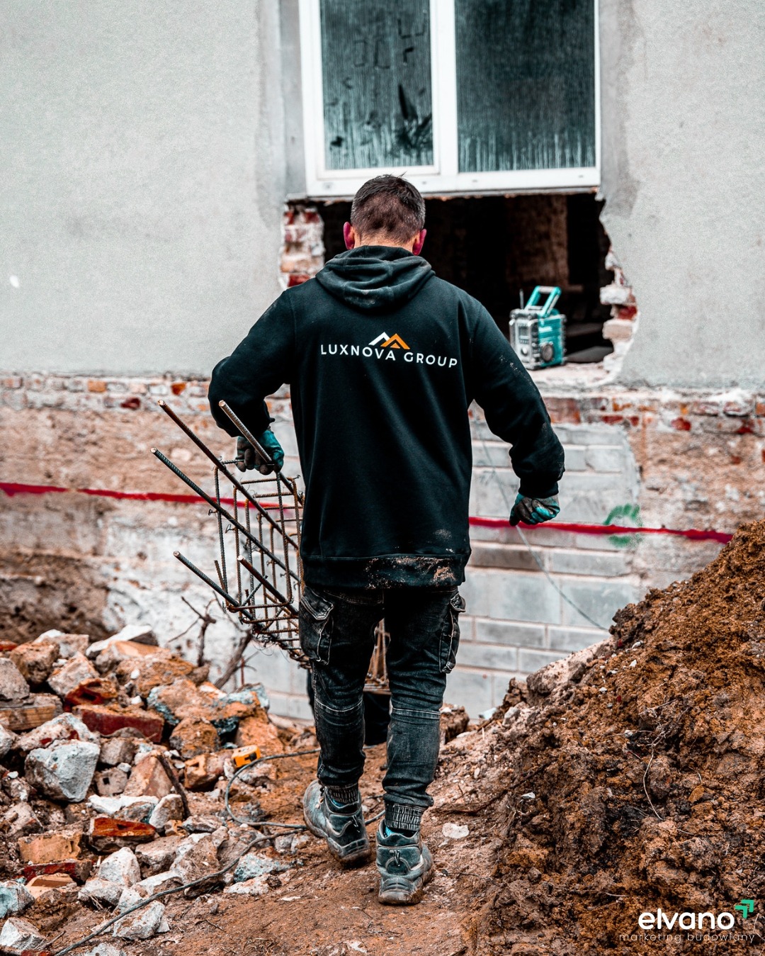 Mężczyzna w czarnej bluzie z logo Luxnova Group, niosący zbrojenie na placu budowy z gruzem i ziemią, w tle uszkodzony budynek z oknem i radiem.
