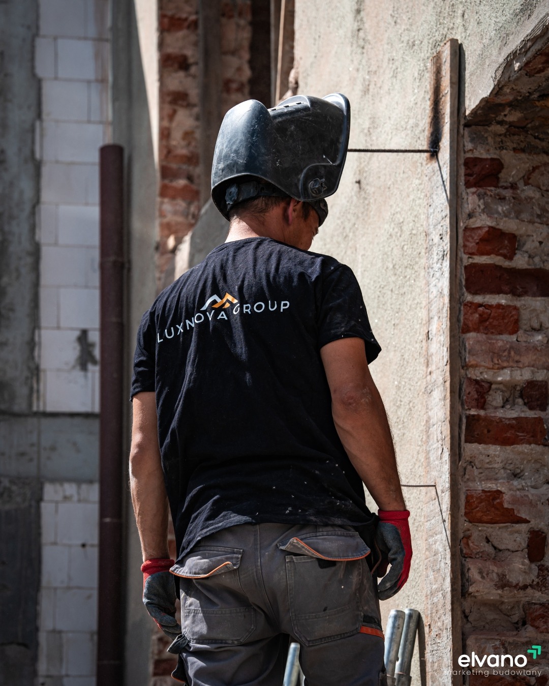 Pracownik w masce spawalniczej, w czarnej koszulce z logo Luxnova Group, wzmacnia ścianę budynku za pomocą metalowych prętów.