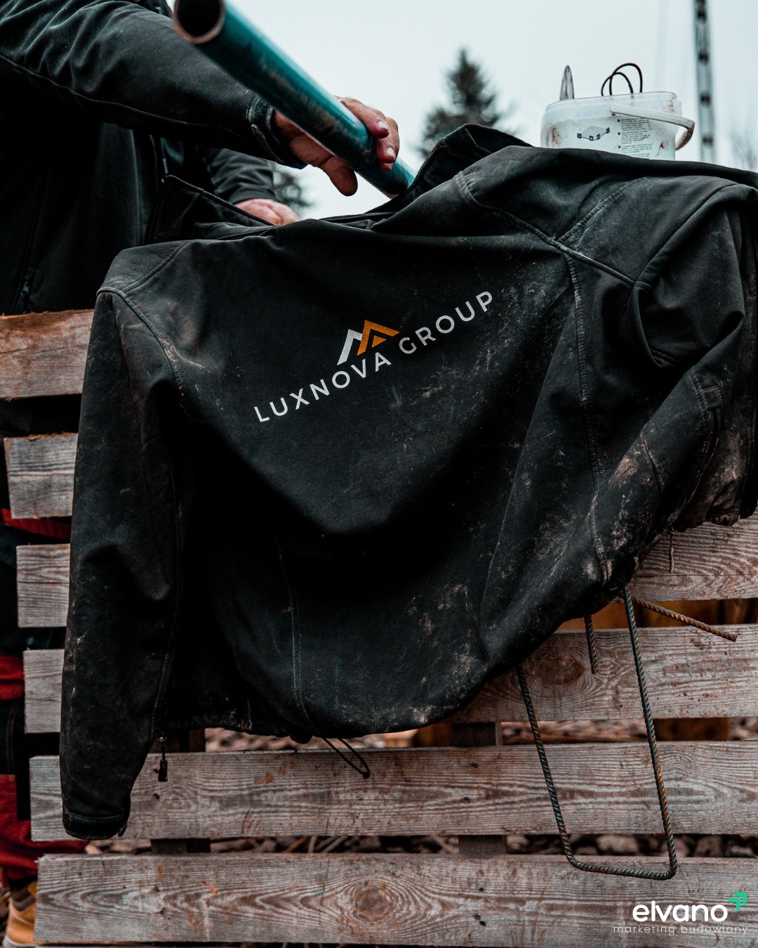 Czarna kurtka robocza z logo 'LUXNOVA GROUP' leżąca na drewnianej palecie, z widocznymi śladami użytkowania i ręką trzymającą niebieską rurę w tle.