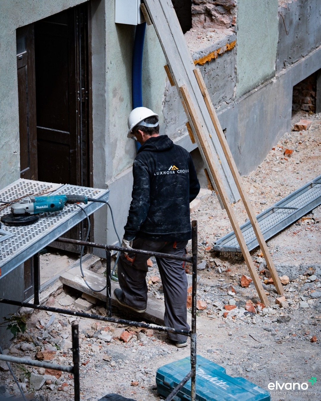 Pracownik w kasku i bluzie z logo firmy Luxnova Group, w trakcie prac remontowych przy budynku, widoczny ruszt, skrzynka narzędziowa i szlifierka kątowa.