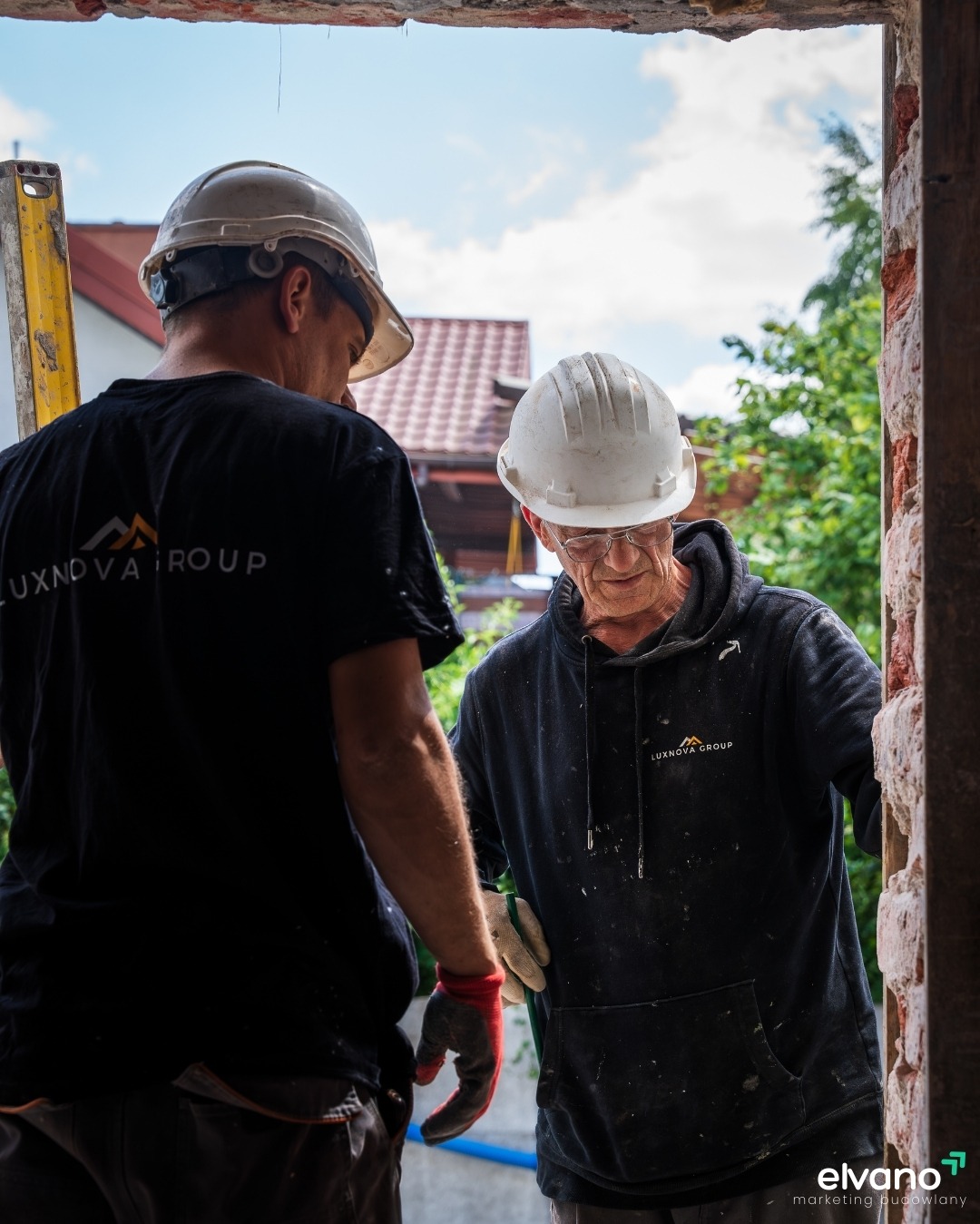 Dwóch pracowników budowlanych w kaskach, jeden w czarnej koszulce z logo, drugi w czarnej bluzie z kapturem, pracują przy murze z cegieł w trakcie remontu okna.