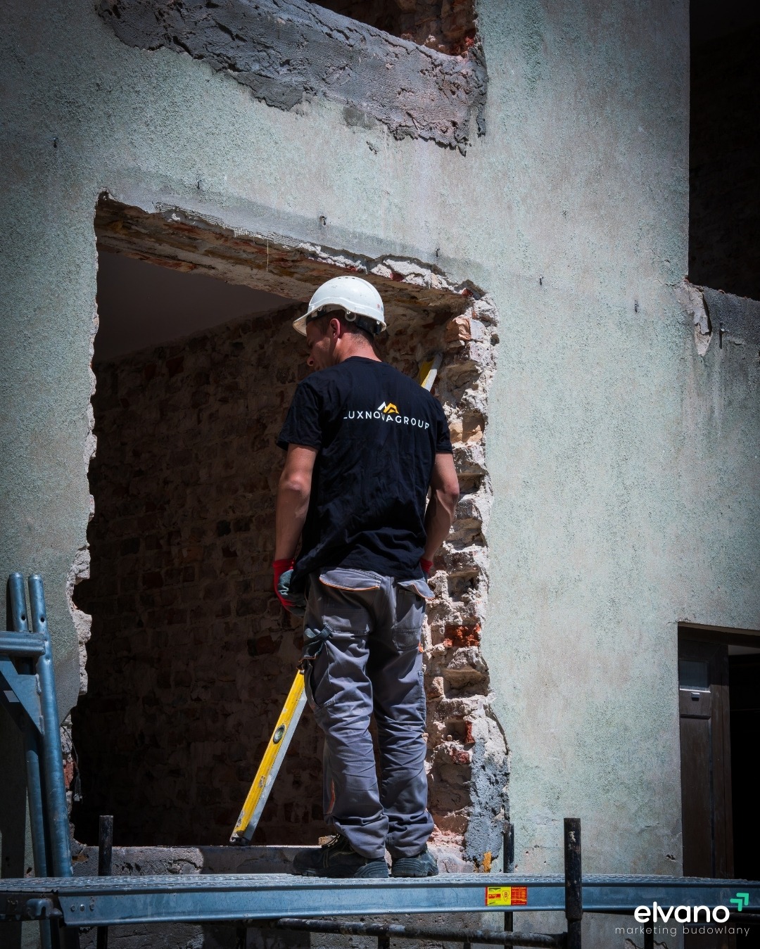 Pracownik budowlany w kasku stoi na rusztowaniu przed surowym otworem w ścianie z cegły, trzymając poziomicę.