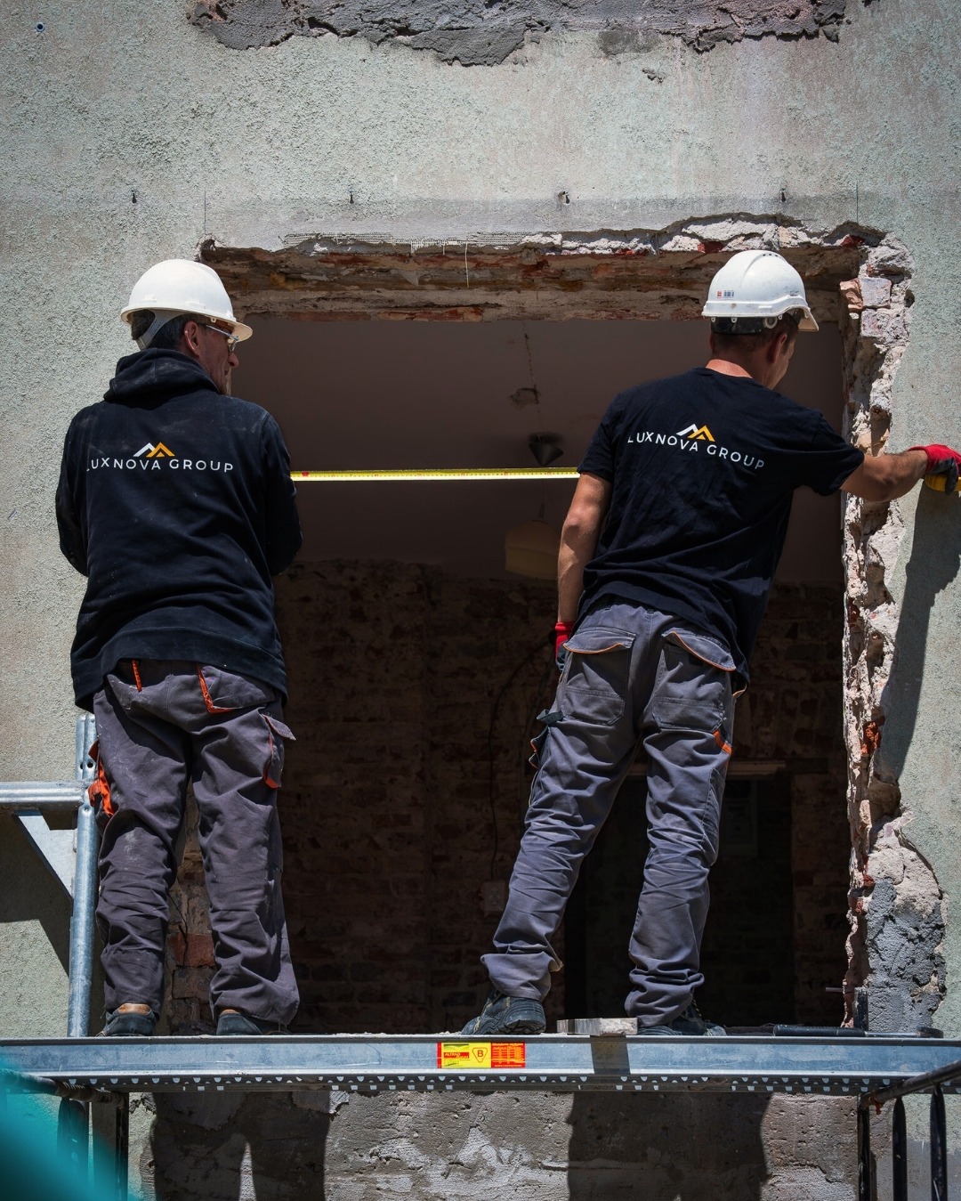 Dwóch pracowników w kaskach i uniformach Luxnova Group mierzy otwór w ścianie z cegły, stojąc na rusztowaniu.