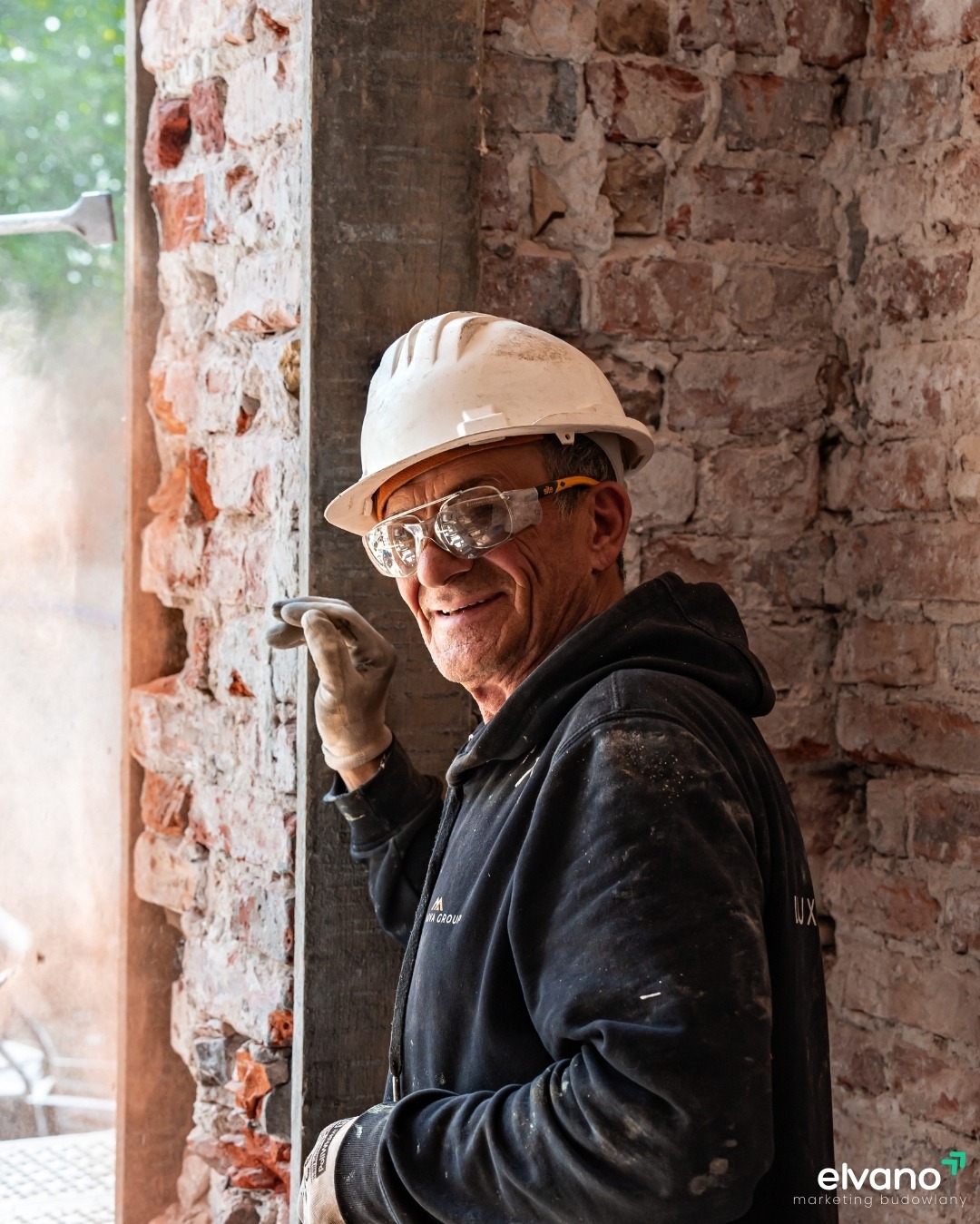 Uśmiechnięty murarz w kasku i okularach ochronnych, opierający się o odrzwia z odzyskaną cegłą, w tle narzędzie pneumatyczne i pył.