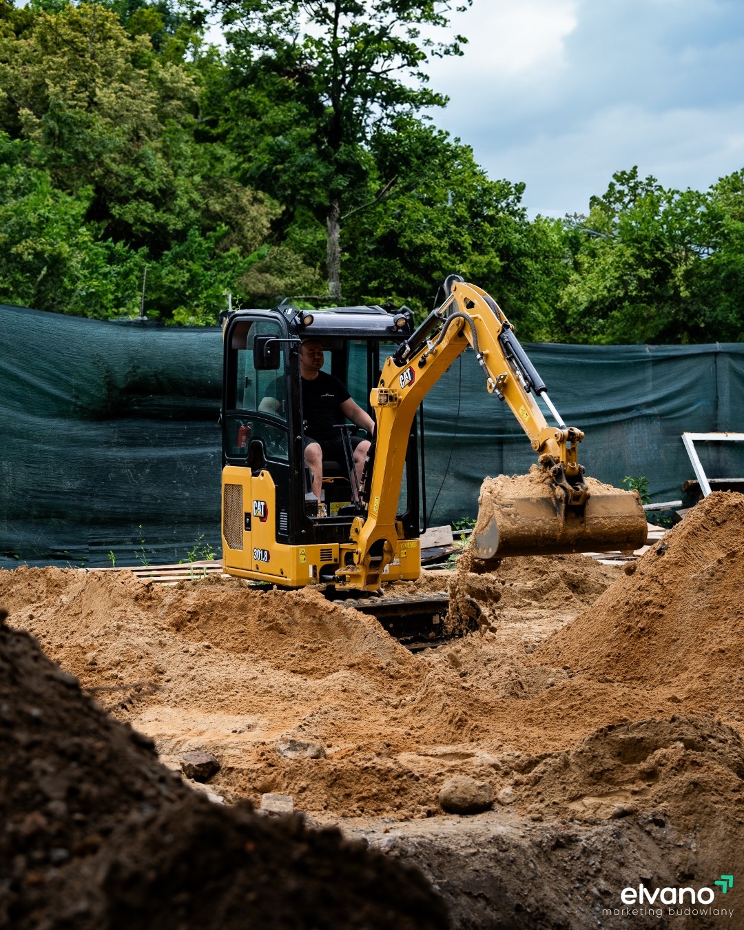 Operator w żółtej minikoparce CAT 301.8, nabierający piasek z wykopu na tle zieleni i ogrodzenia, z logo firmy budowlanej w prawym dolnym rogu.