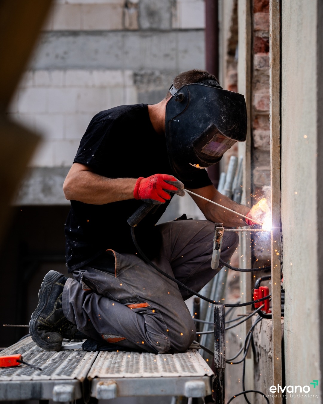 Spawacz w masce i czerwonych rękawicach klęczy na rusztowaniu podczas spawania metalowej konstrukcji, iskry rozbryzgują się na boki, w tle surowe ściany budynku w trakcie budowy.