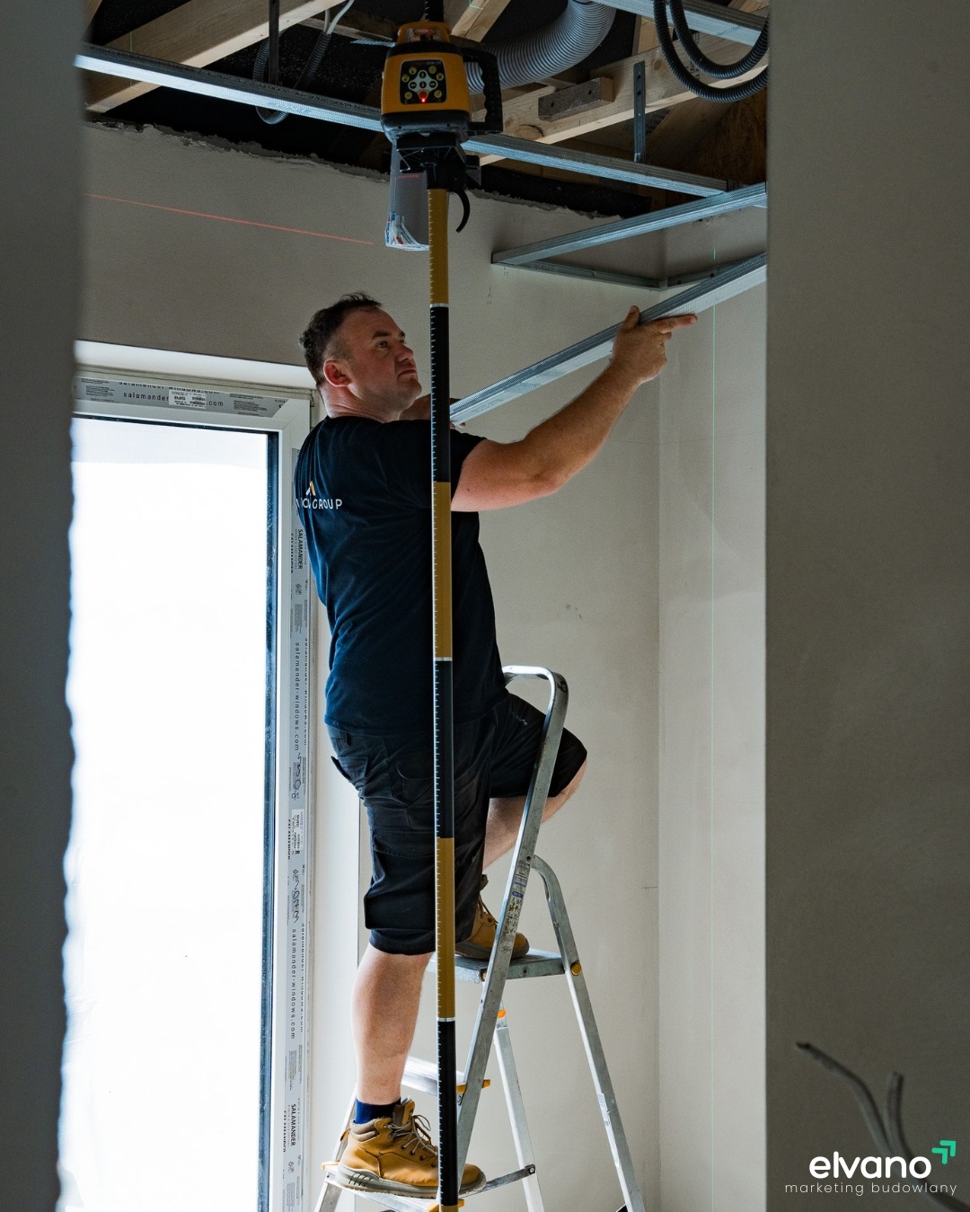 Mężczyzna na drabinie montuje metalowy stelaż sufitu podwieszanego, używając poziomicy laserowej z czerwoną wiązką, widoczne narzędzia i elementy konstrukcyjne.