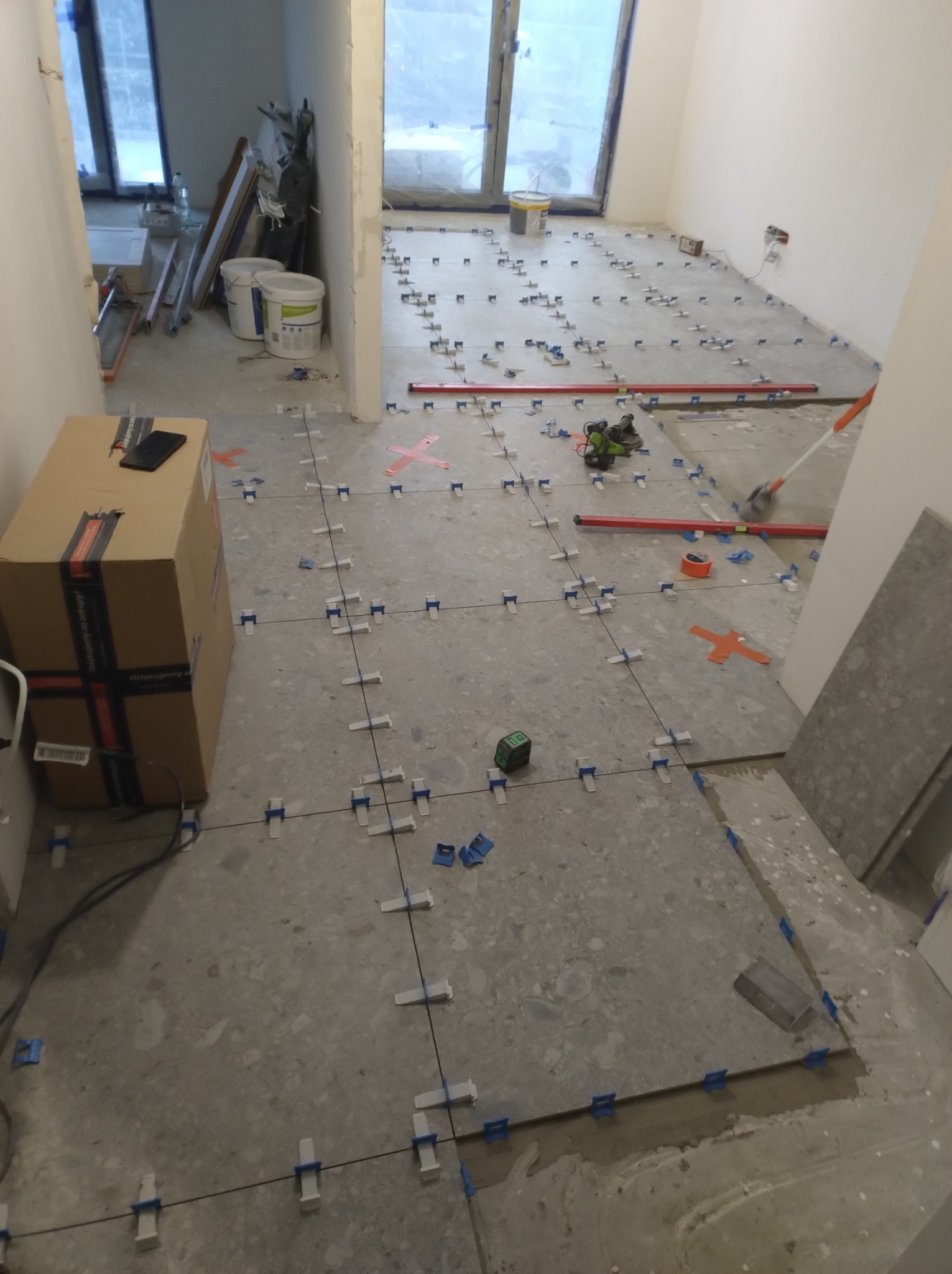 Układanie płytek podłogowych w trakcie remontu mieszkania, widoczne krzyżyki dystansowe, poziomica i laserowy miernik. Częściowo ułożona podłoga w szarym kolorze.
