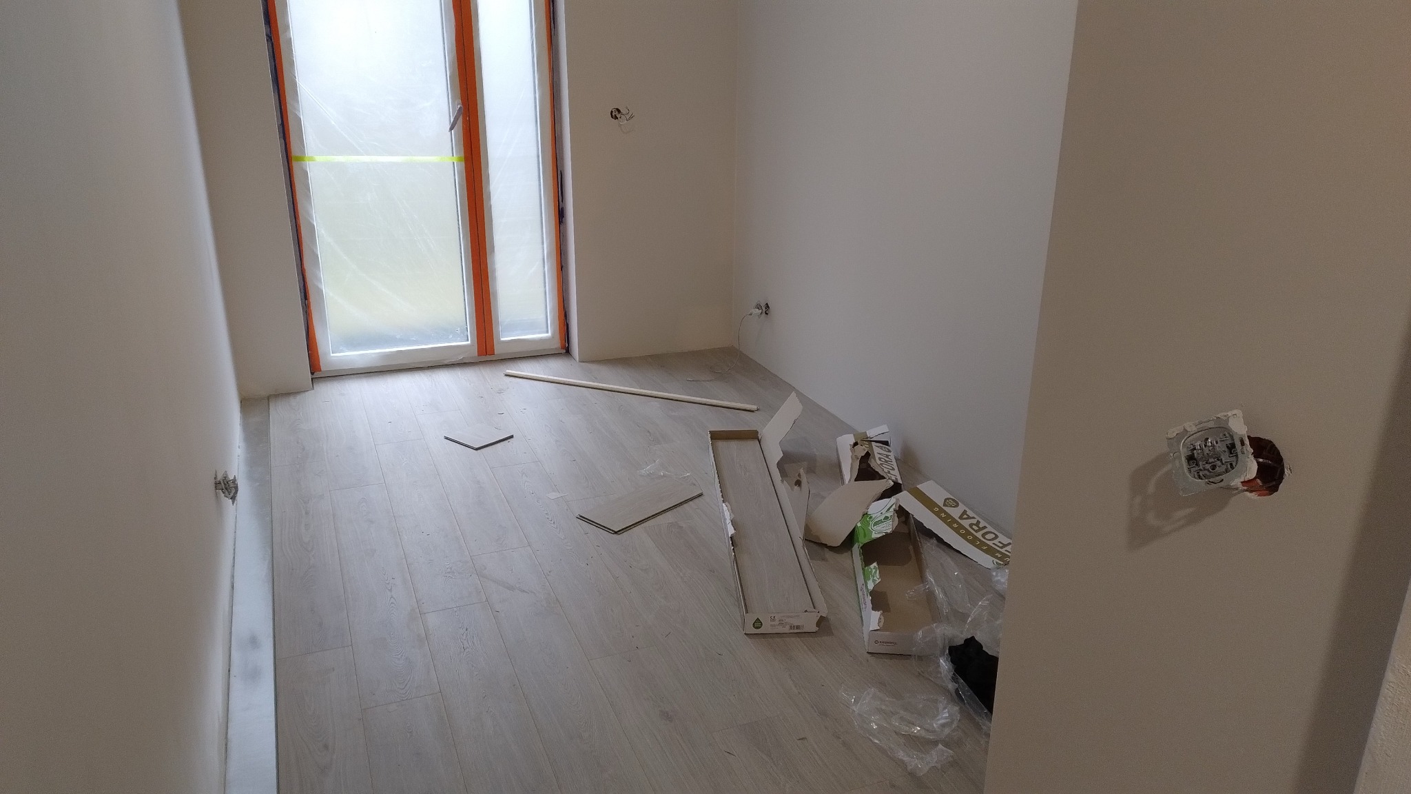 Układanie paneli podłogowych w jasnym pokoju. Widoczne otwarte pudełka z panelami, listwa przypodłogowa i gniazdko elektryczne w ścianie.