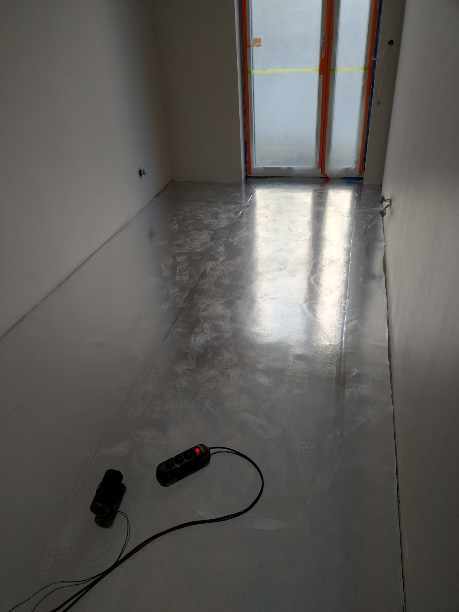 Błyszcząca, szara wylewka samopoziomująca na podłodze w trakcie remontu mieszkania. Widoczne ślady stóp i przewody elektryczne.