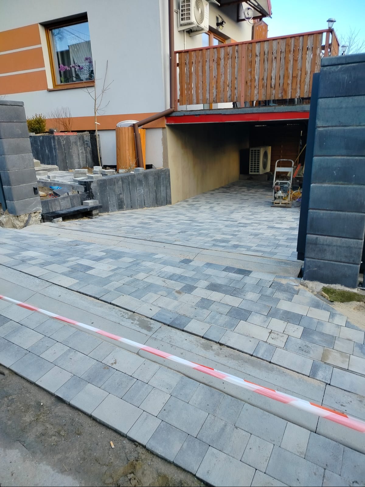 Układanie kostki brukowej na podjeździe do garażu. Widoczna maszyna do ubijania, fragment domu z klimatyzacją i drewnianą barierką. Częściowo ułożona kostka w odcieniach szarości.