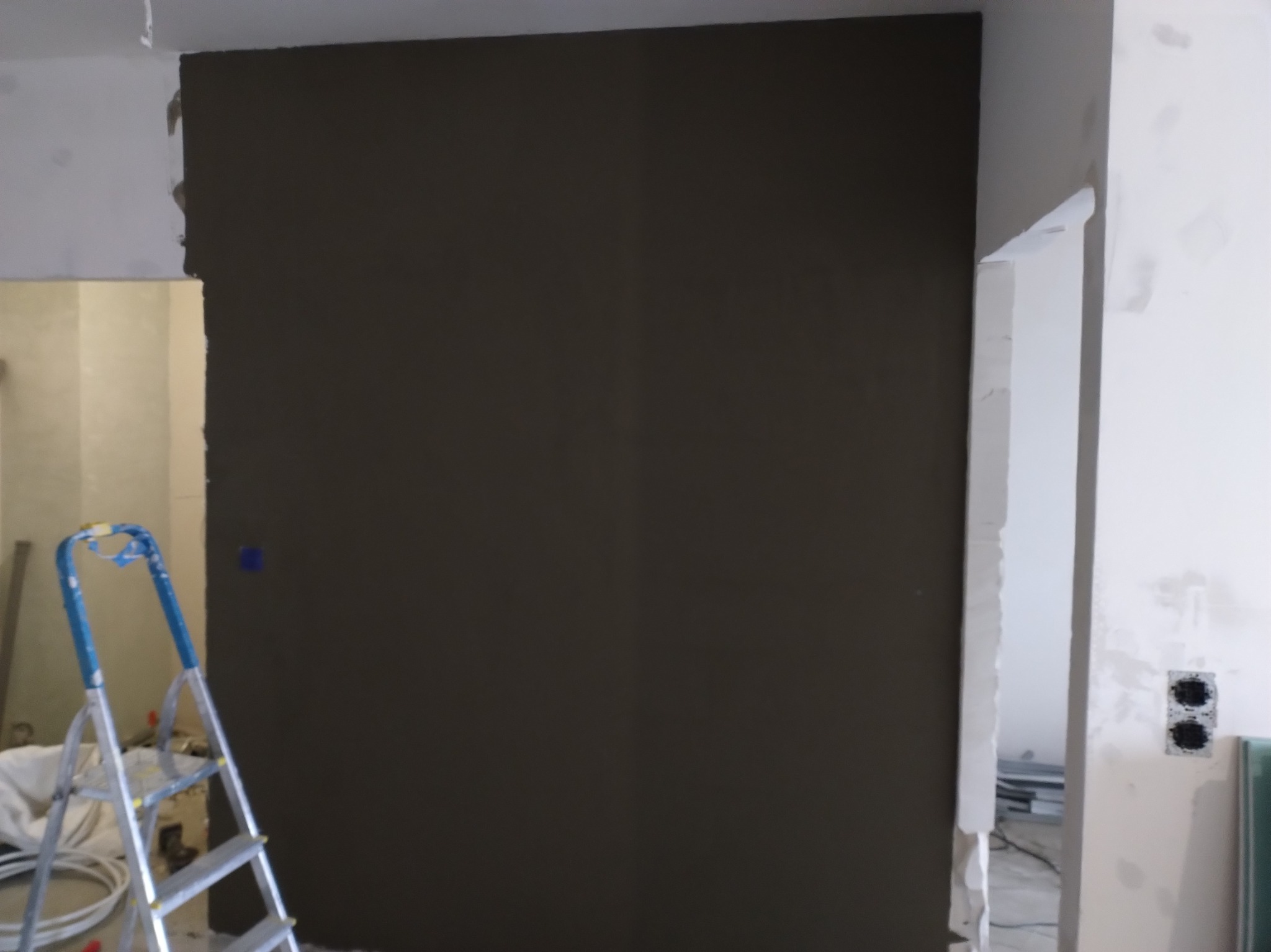 Ściana po nałożeniu ciemnego tynku, widoczna drabina malarska i przygotowane gniazdka elektryczne. Etap prac wykończeniowych w mieszkaniu.
