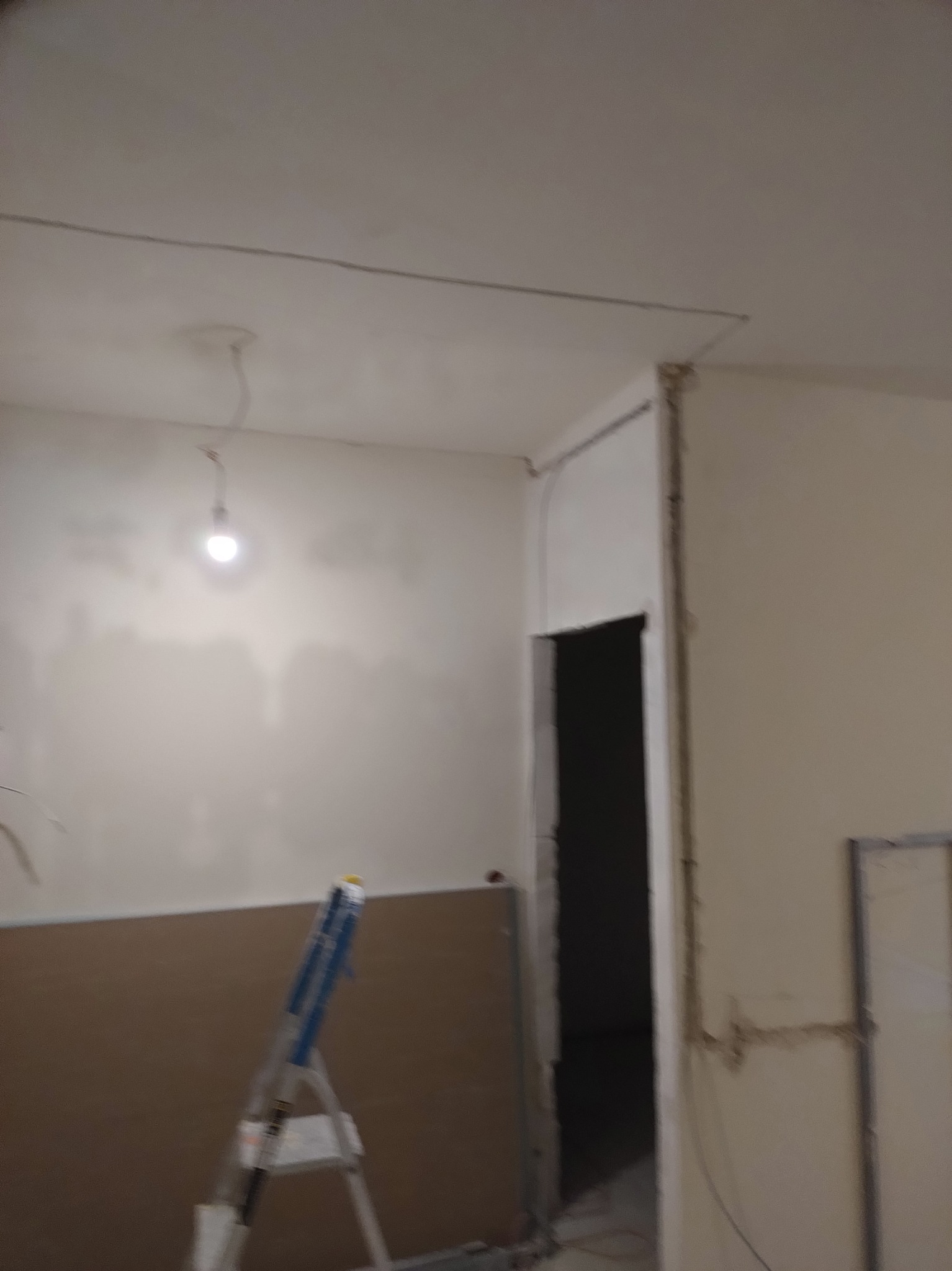 Wnętrze w trakcie remontu: widoczna drabina, ściany przygotowane do malowania, odsłonięte kable elektryczne i surowy otwór drzwiowy. Praca w toku.