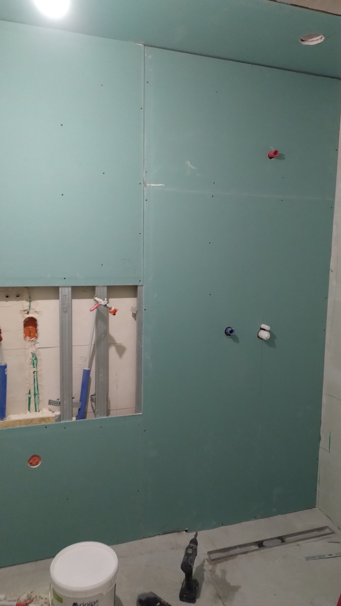 Montaż płyt gipsowo-kartonowych w łazience, widoczne instalacje wodne i elektryczne. Na podłodze wiadro z farbą i wkrętarka.