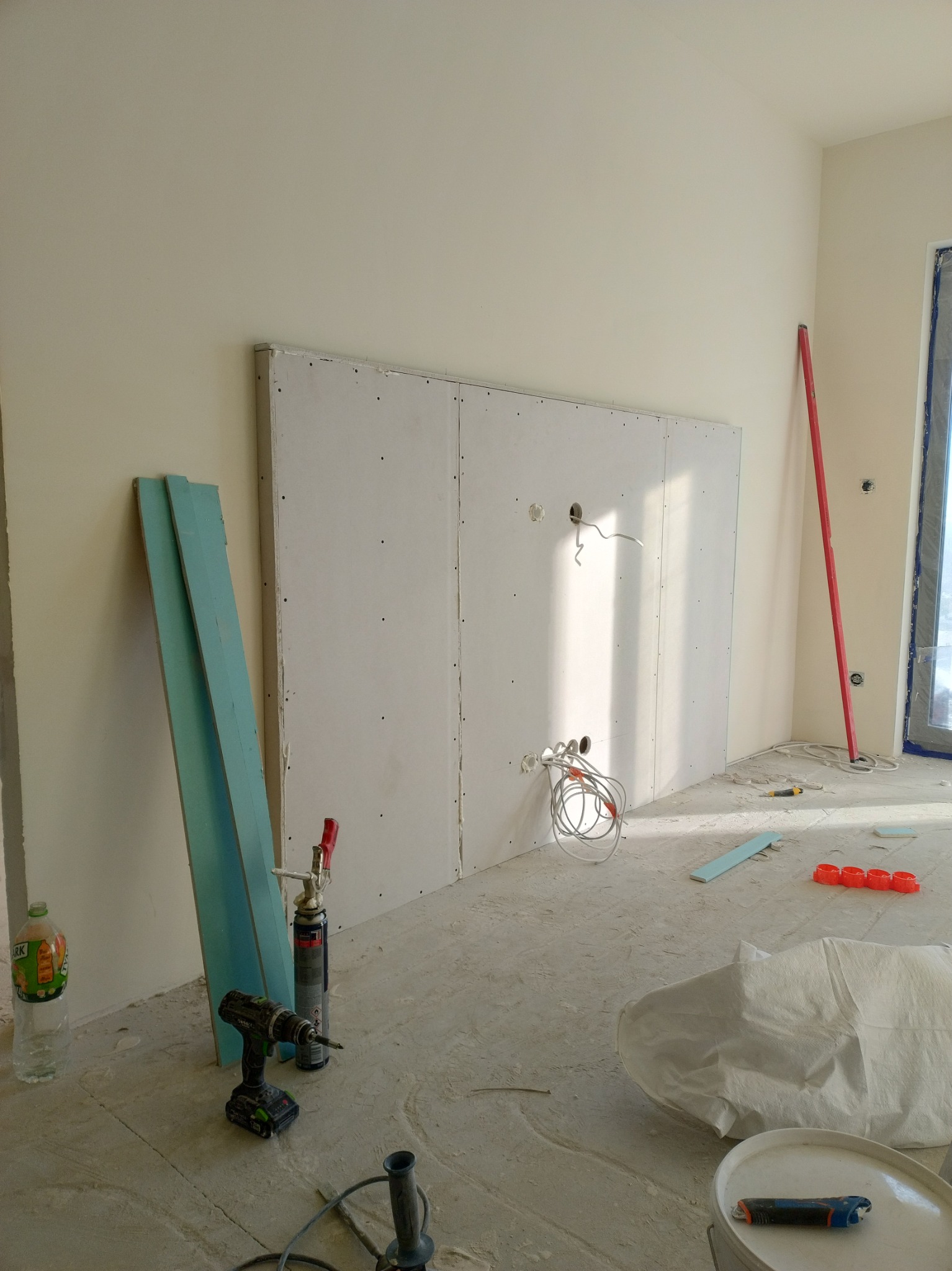 Montaż płyty gipsowej na ścianie z widocznymi przewodami elektrycznymi, narzędziami i materiałami budowlanymi w trakcie remontu mieszkania.