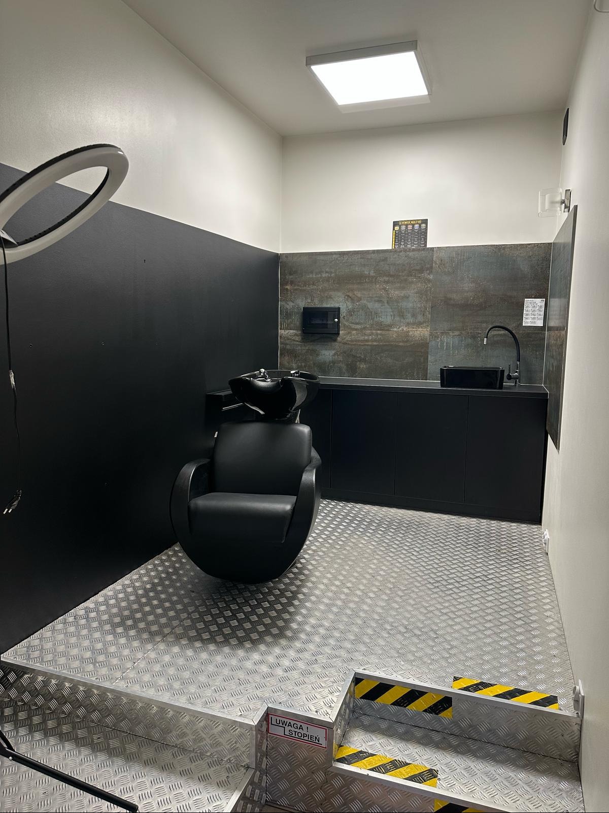 Nowoczesna aranżacja wnętrza salonu fryzjerskiego: czarne meble, fotel z myjnią, metalowa podłoga z ostrzegawczymi pasami. Minimalizm i funkcjonalność.