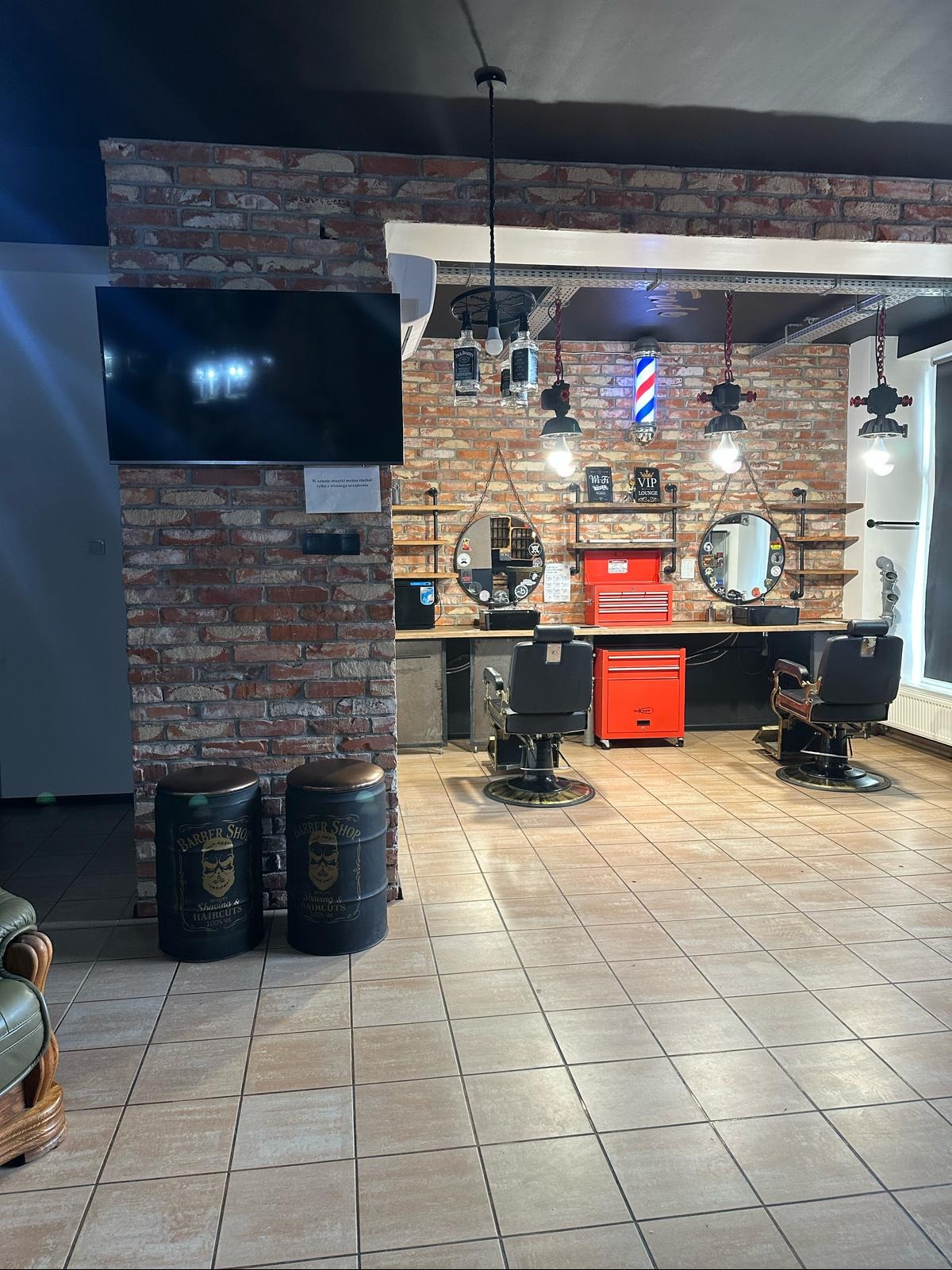 Wnętrze barber shopu z ceglaną ścianą, dwoma fotelami fryzjerskimi, narzędziami i dekoracjami w stylu vintage, z widocznymi beczkami z napisem Barber Shop.