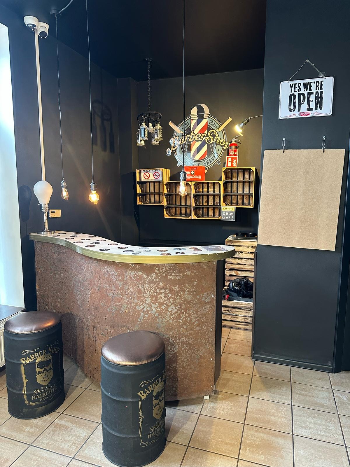 Wnętrze barber shopu: rustykalna lada z dekoracyjnymi krzesłami-beczkami, oświetlenie w stylu retro i czarne ściany z półkami z drewnianych skrzynek.