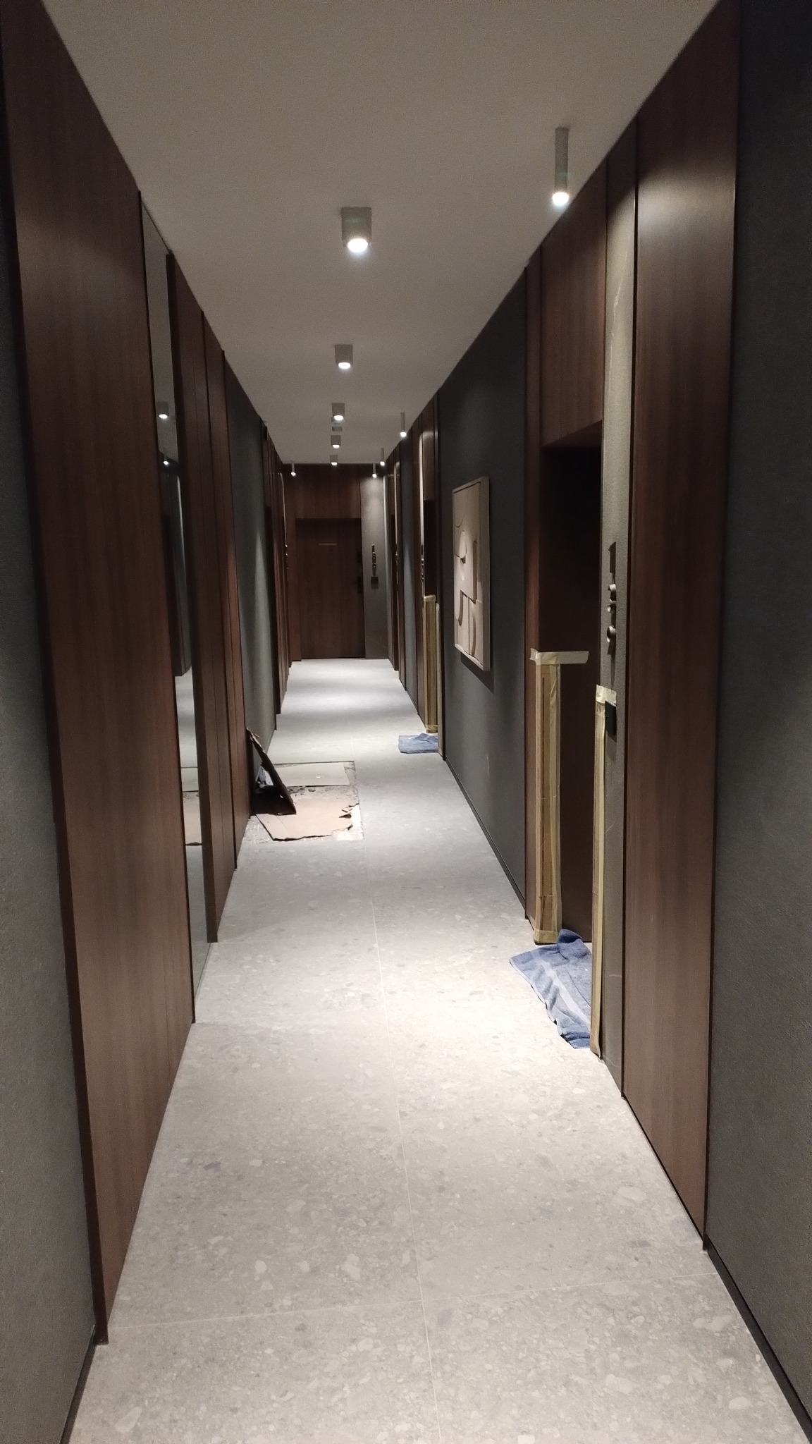 Długi korytarz po remoncie z nową, jasną podłogą z płytek. Ściany wykończone w drewnie i szarości. Widoczne zabezpieczenia na drzwiach i fragmenty prac remontowych.