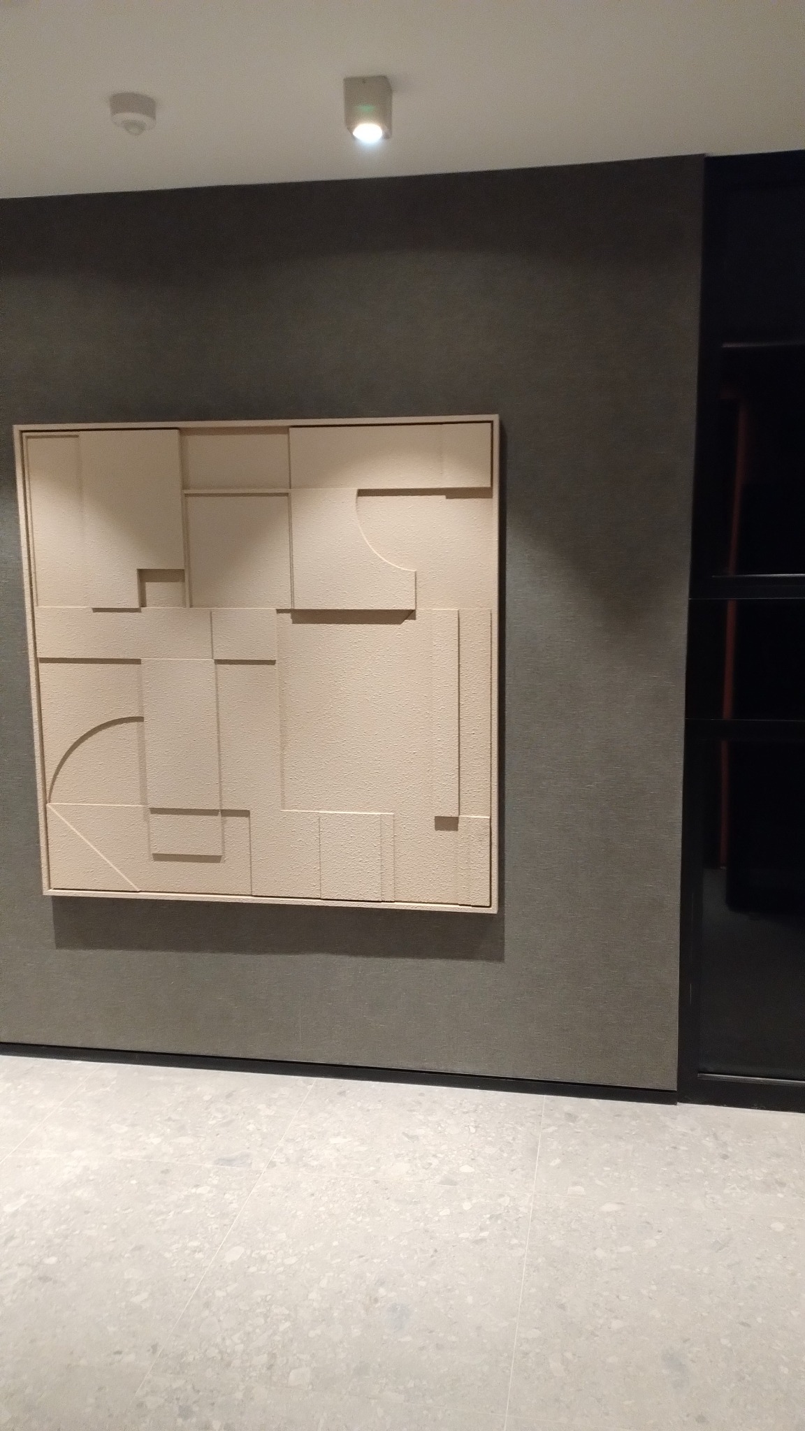 Abstrakcyjna płaskorzeźba na ścianie w Tychach. Geometryczne formy w odcieniach beżu, oświetlenie sufitowe. Minimalistyczny design wnętrza.