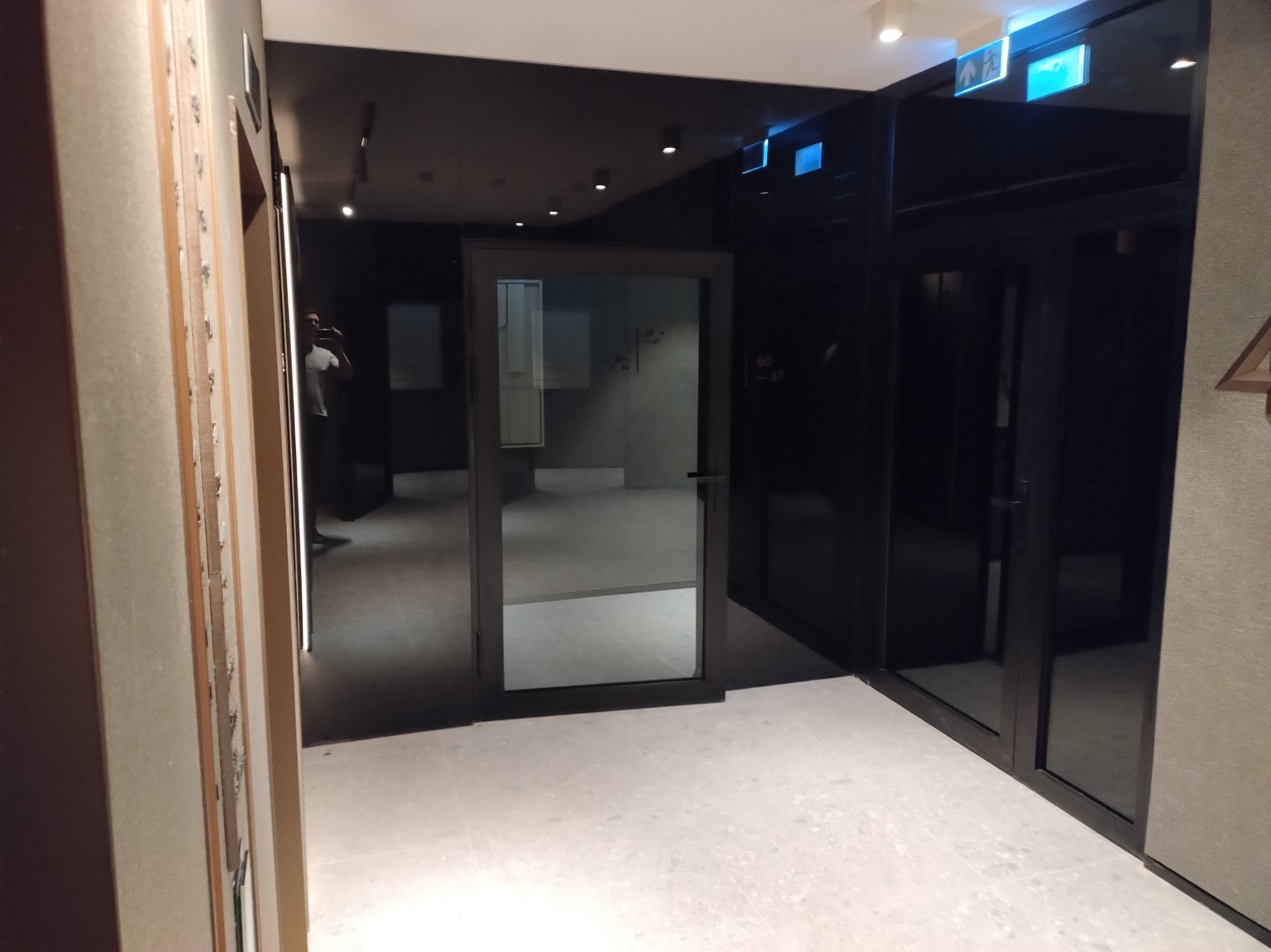 Nowoczesne wnętrze z czarnymi ścianami i szklanymi drzwiami. Widoczne oświetlenie sufitowe i jasna podłoga. Fragment ściany z numerem apartamentu.