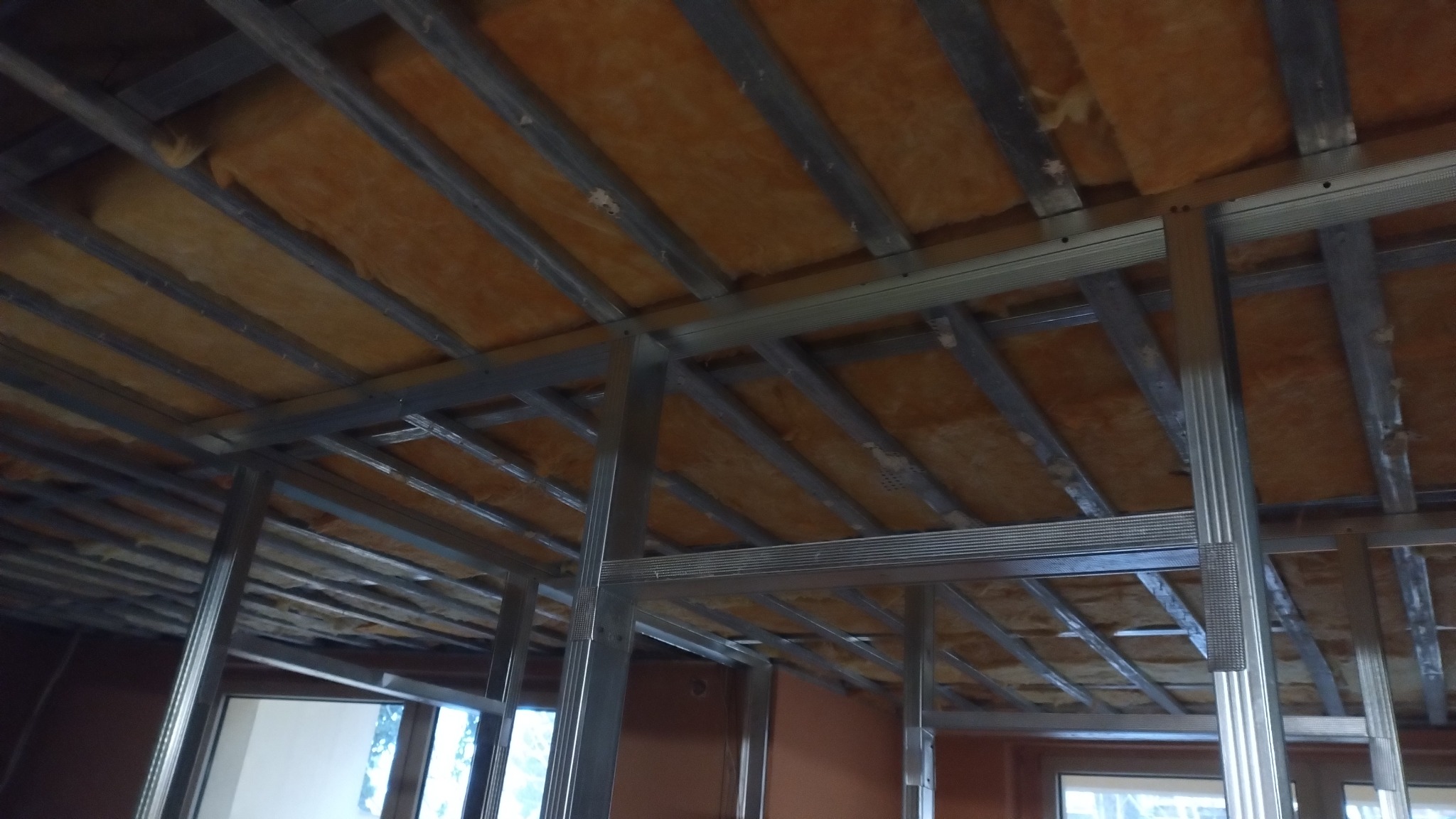 Montaż izolacji z wełny mineralnej w metalowej konstrukcji sufitu podwieszanego, widok z dołu, etap prac wykończeniowych wewnątrz remontowanego mieszkania.