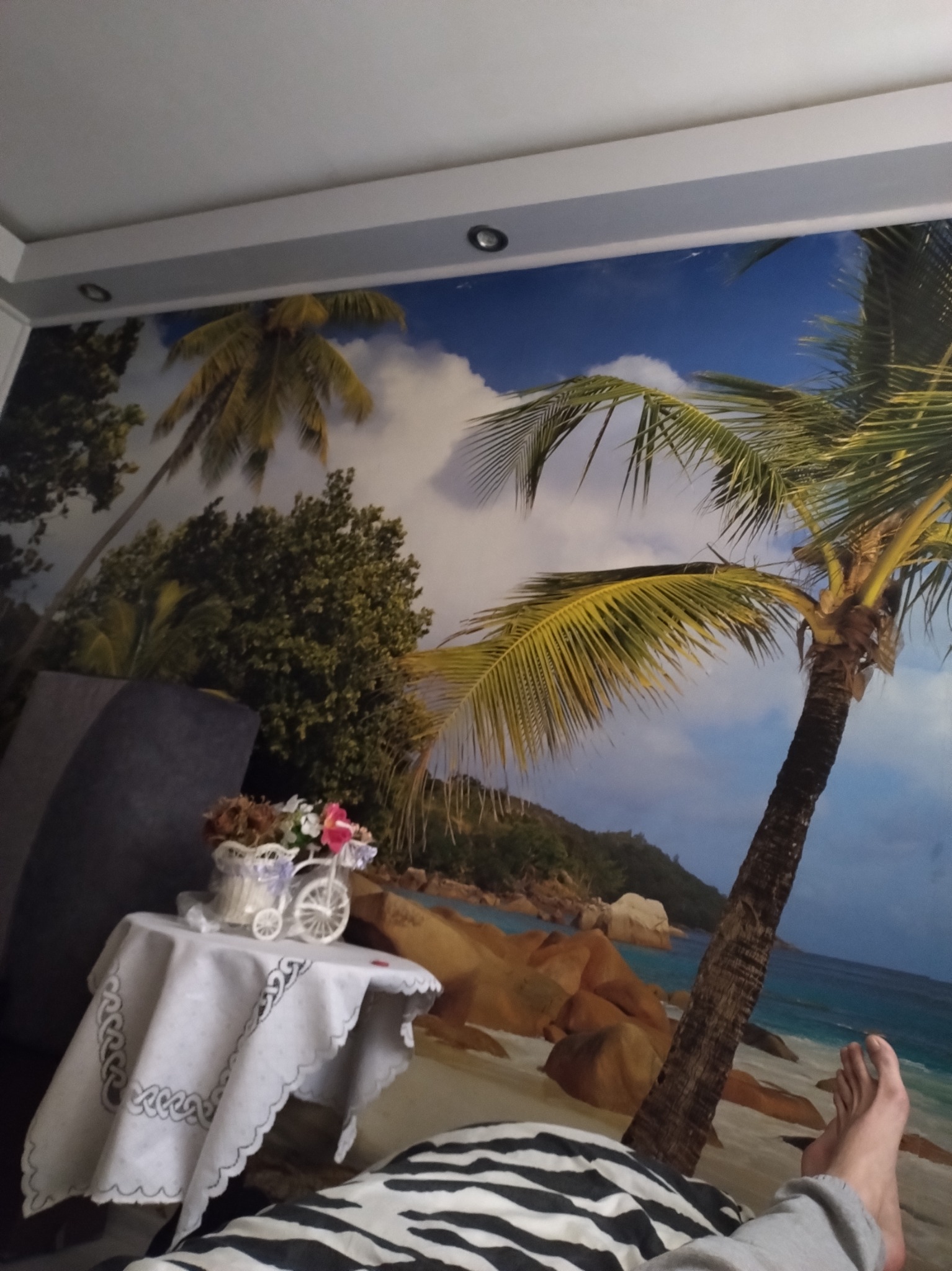 Fototapeta z motywem tropikalnej plaży w pokoju po remoncie. Na pierwszym planie stolik z dekoracją kwiatową i fragment leżącej osoby.