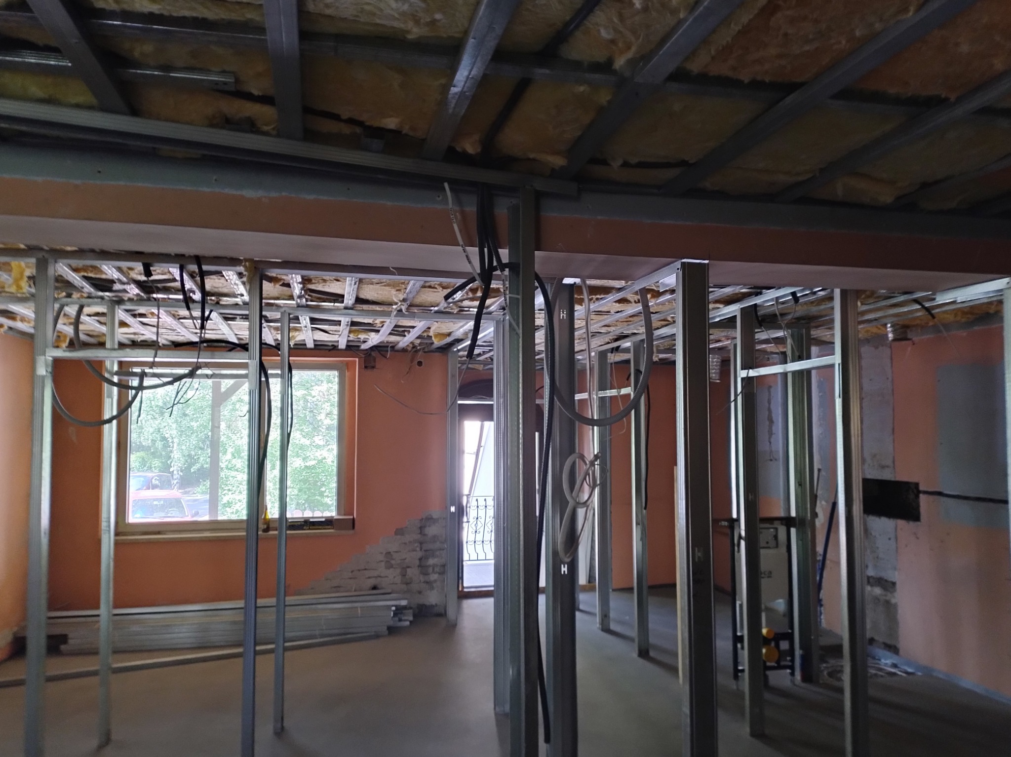 Wnętrze w trakcie remontu: metalowa konstrukcja ścian działowych, izolacja sufitu, instalacje elektryczne. Widoczne okno i fragment ściany z odsłoniętą cegłą.