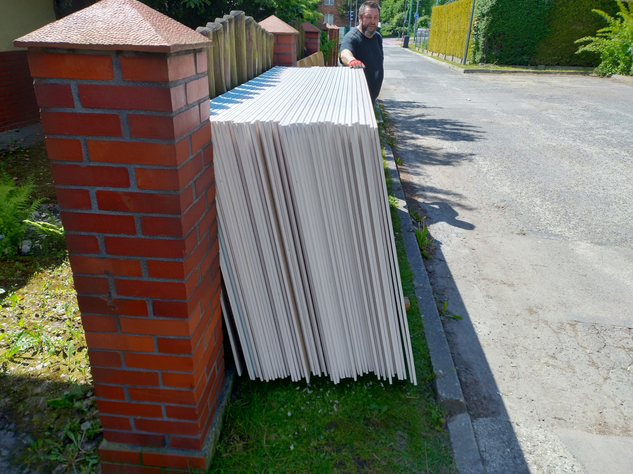 Stos białych płyt gipsowo-kartonowych oparty o ceglany filar ogrodzenia, przygotowany do montażu wewnątrz budynku. Mężczyzna w tle nadzoruje transport.