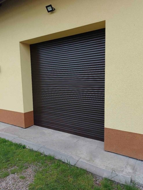 Zamknięta, brązowa roleta garażowa w elewacji budynku z lampą nad nią. Widoczny cokół i fragment trawnika.