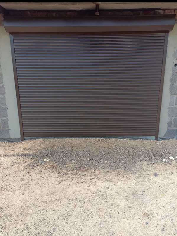 Zamknięta, brązowa brama rolowana garażu z widoczną fakturą blachy, zamontowana w surowym murze z betonu komórkowego. Podłoże: żwir.