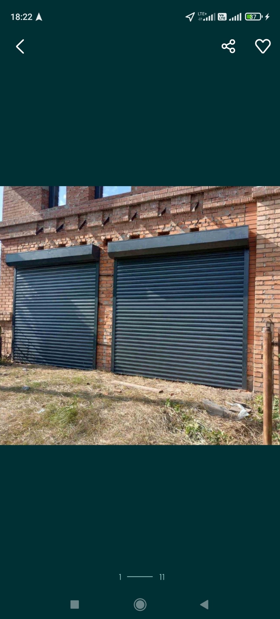 Dwie zamknięte, czarne bramy rolowane z poziomymi panelami, zamontowane w ceglanym budynku. Widoczne trawa i ziemia przed budynkiem.