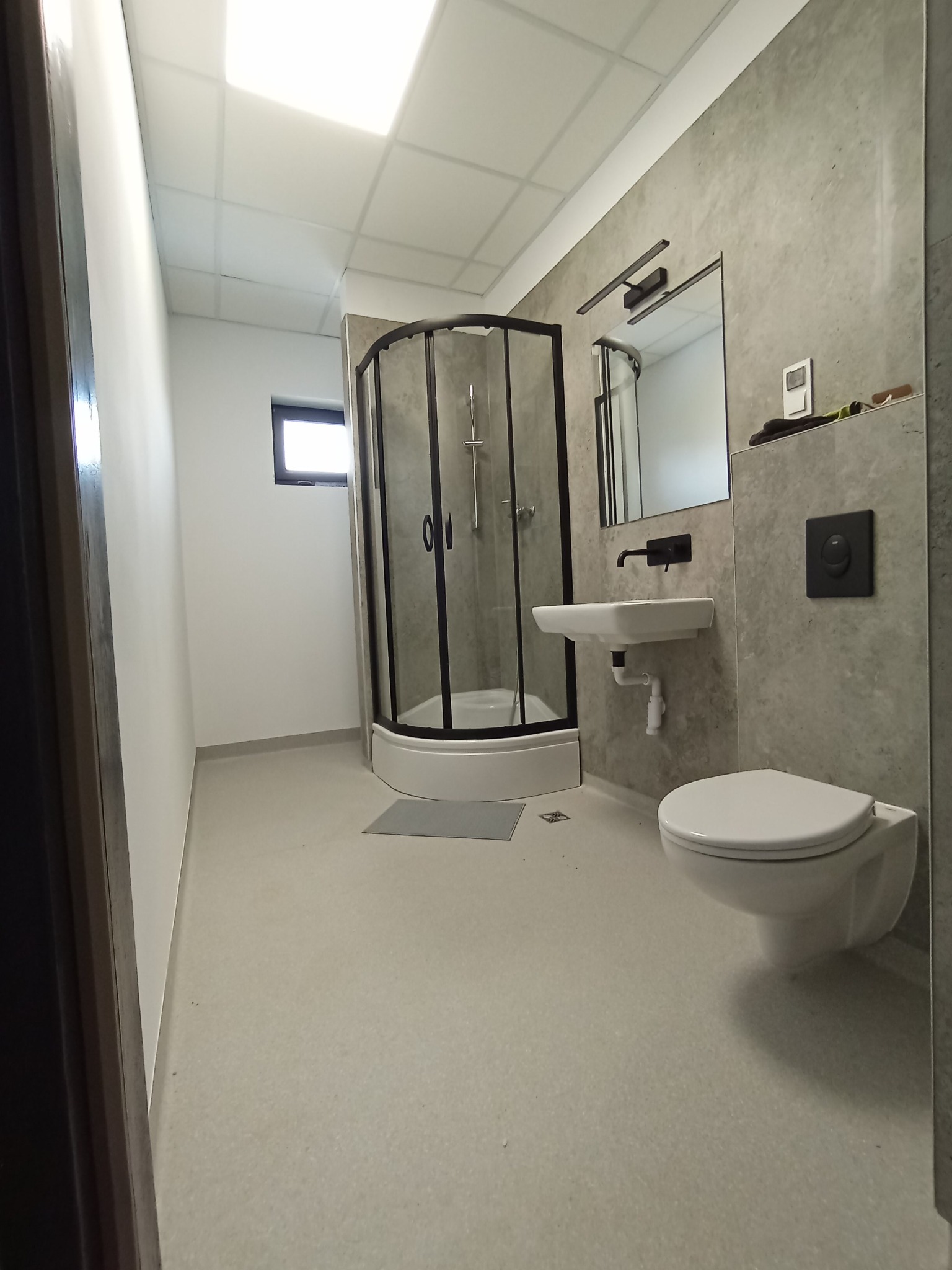 Nowoczesna łazienka z kabiną prysznicową w rogu, umywalką na ścianie i podwieszaną toaletą. Szare ściany imitujące beton, czarne ramy kabiny i armatura.