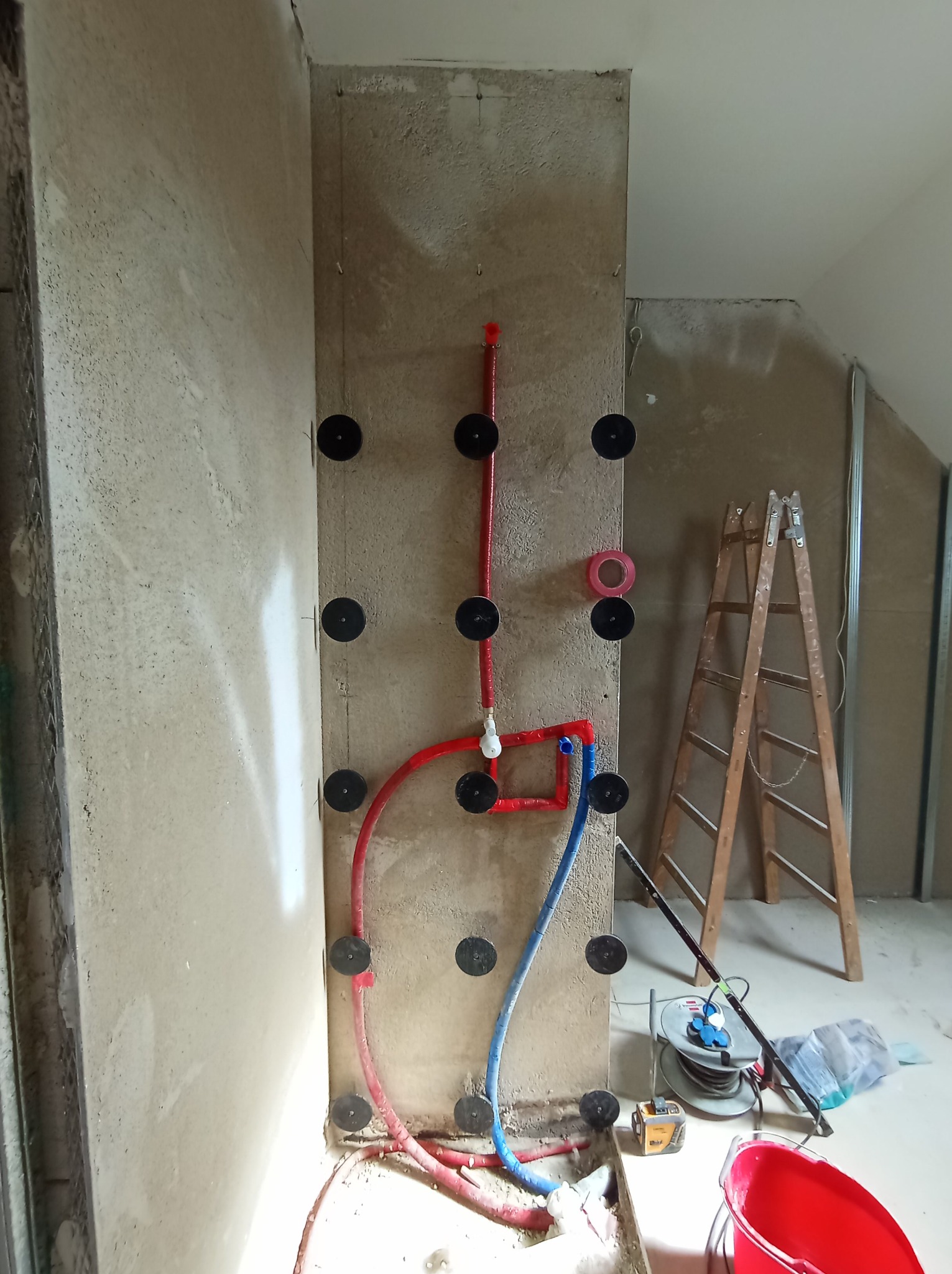 Instalacja wodna w trakcie montażu na ścianie w surowym stanie, widoczne rury w kolorze czerwonym i niebieskim, przymocowane do ściany za pomocą uchwytów.