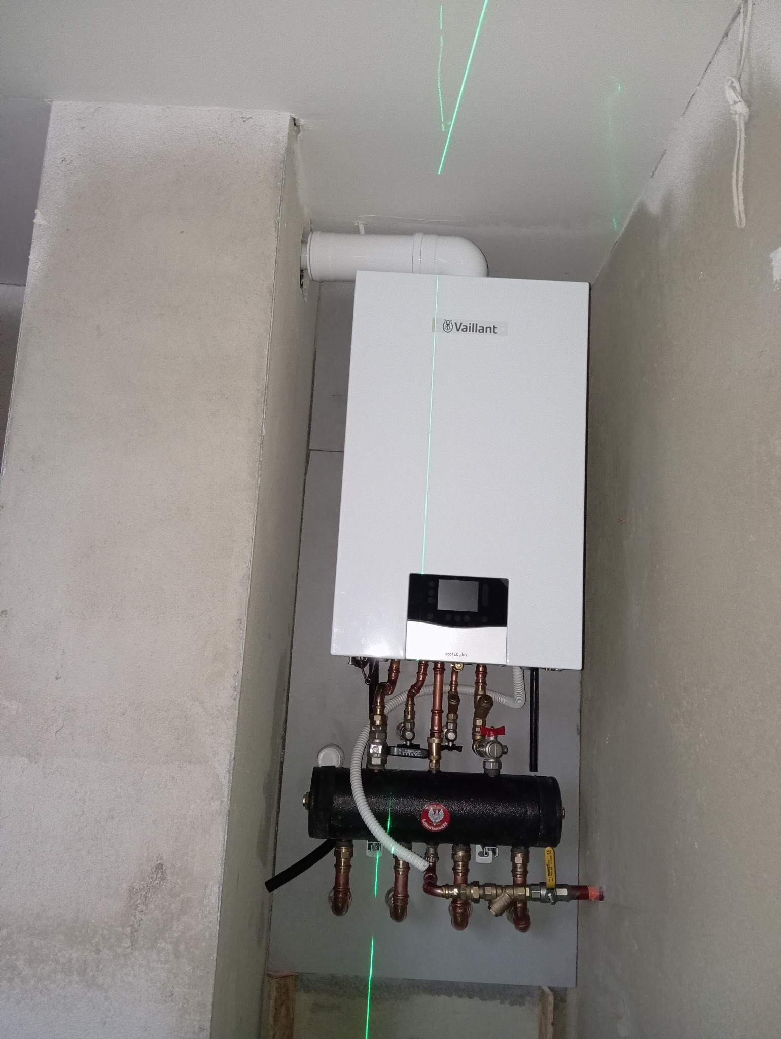 Biały piec gazowy Vaillant z podłączeniami miedzianymi i czarnym zbiornikiem, zamontowany na szarej ścianie. Widoczne zielone linie lasera.