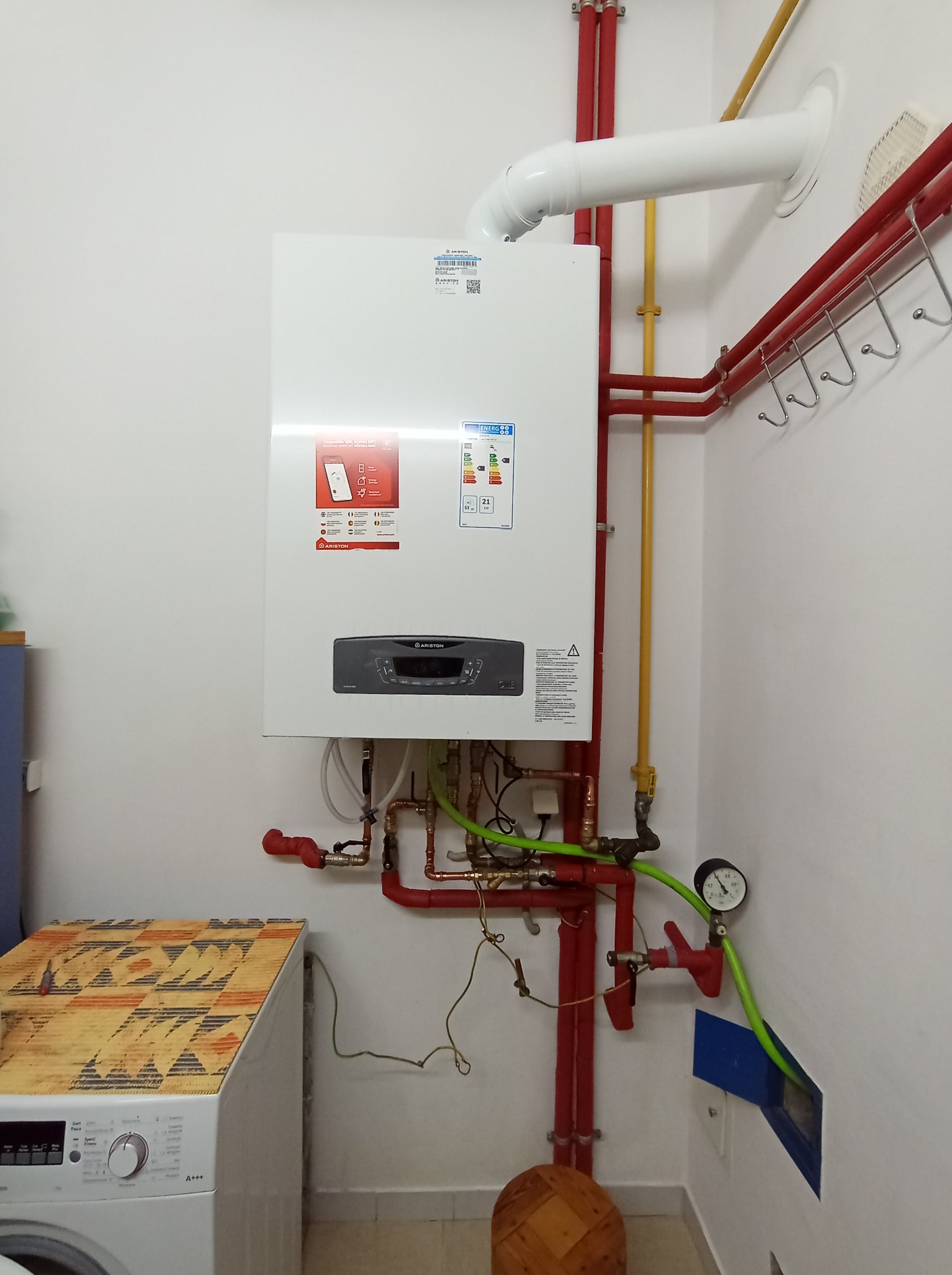 Biały piec gazowy Ariston na ścianie z widoczną instalacją hydrauliczną w kolorze czerwonym i żółtym. W tle pralka z drewnianym blatem i wieszaki na ścianie.