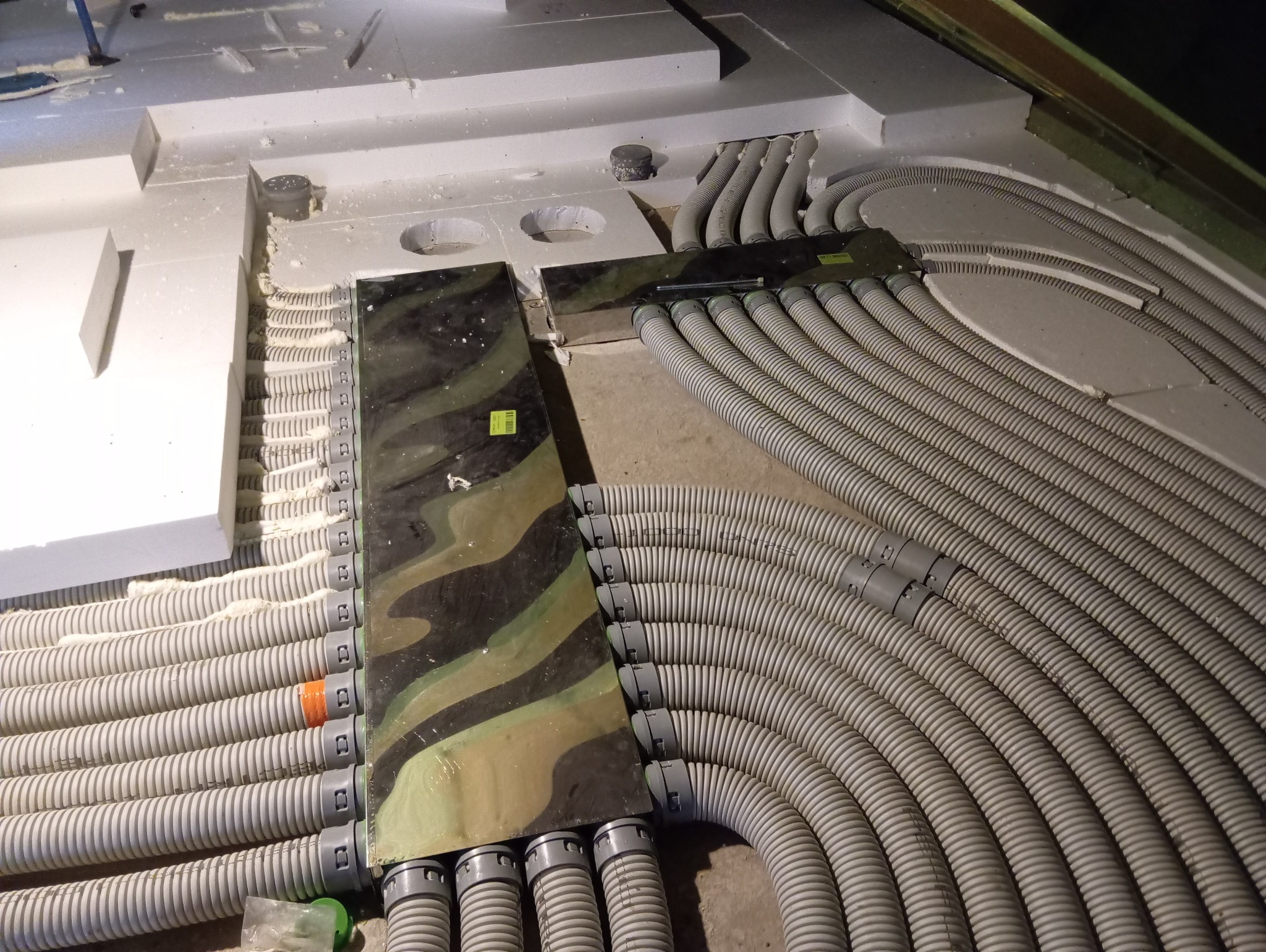 Szare, karbowane rury ogrzewania podłogowego ułożone w izolacji, przymocowane do rozdzielacza z wzorem moro. Widoczne łączenia rur i pianka montażowa.