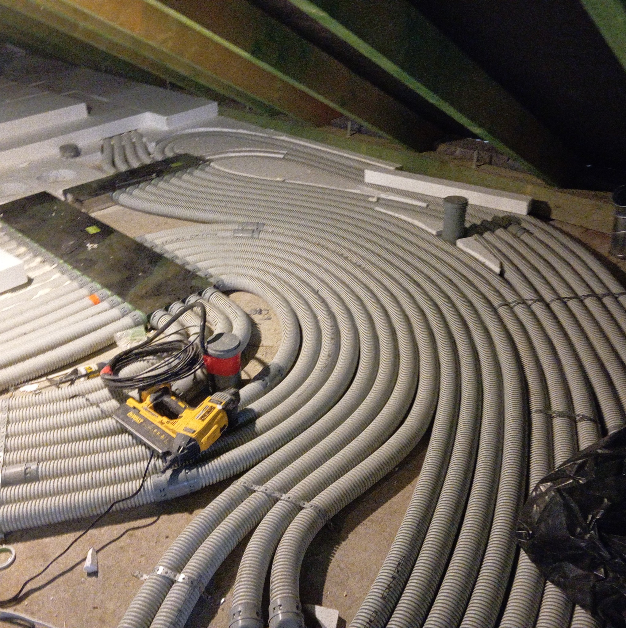 Szare, karbowane rury ogrzewania podłogowego ułożone spiralnie na poddaszu, z narzędziami montażowymi DeWalt obok. Widoczne drewniane elementy konstrukcyjne dachu.