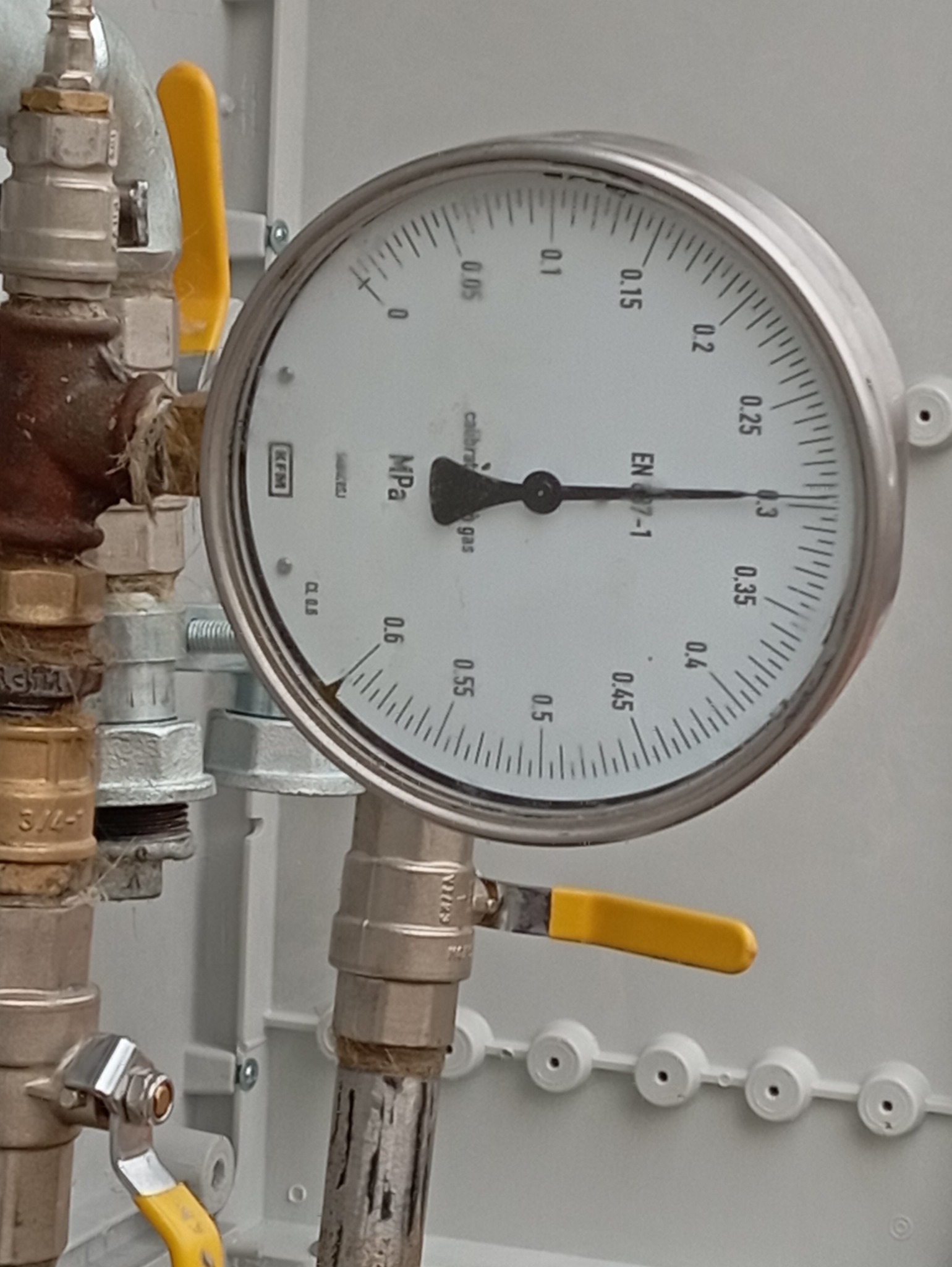 Manometr wskazujący ciśnienie w instalacji gazowej, z widocznymi zaworami i elementami przyłączeniowymi. Układ w szarej obudowie.