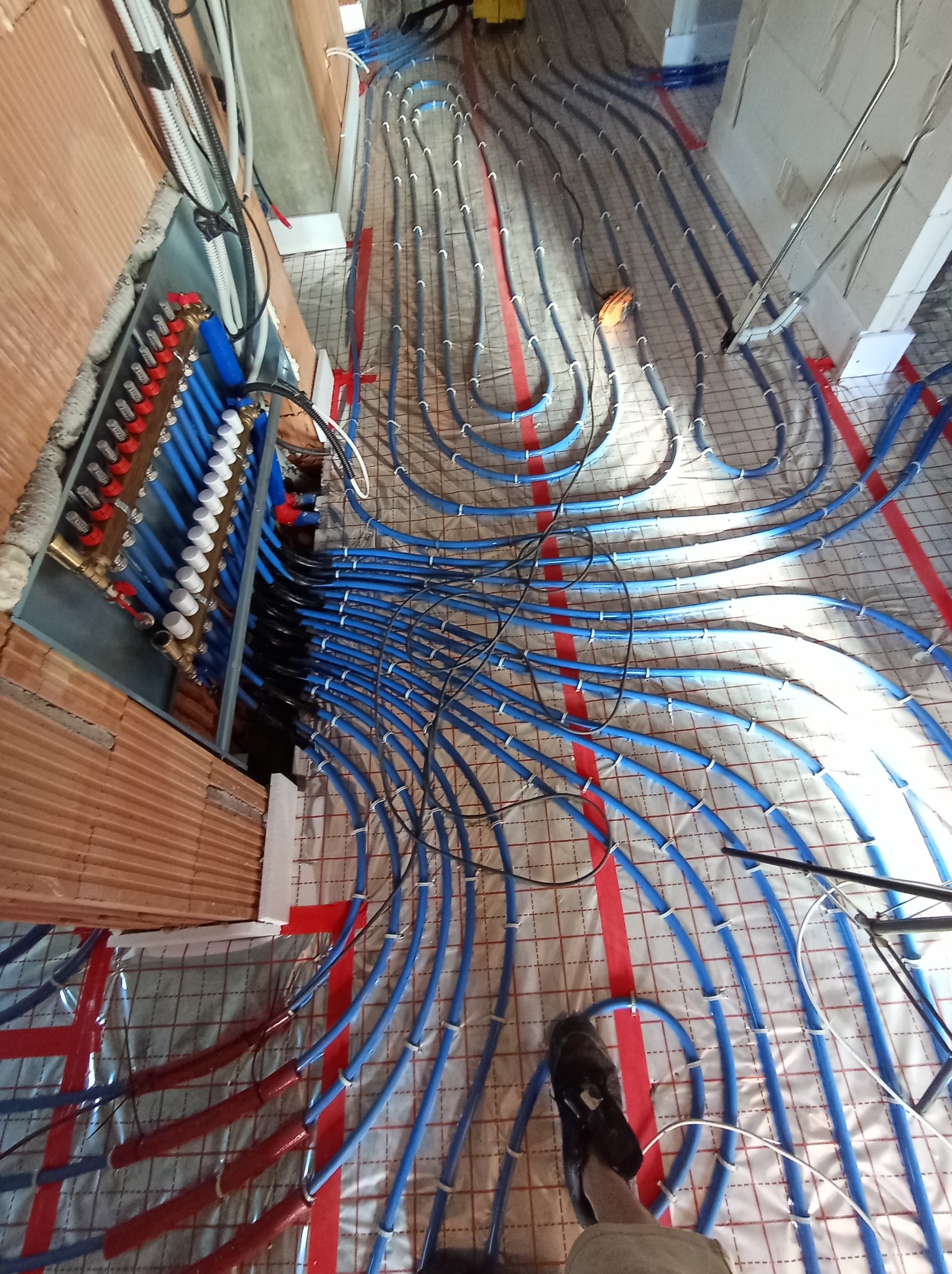 Instalacja ogrzewania podłogowego z rozdzielaczem w Skawinie. Niebieskie rury ułożone na folii z siatką, widoczny rozdzielacz z zaworami i czerwonymi pokrętłami.