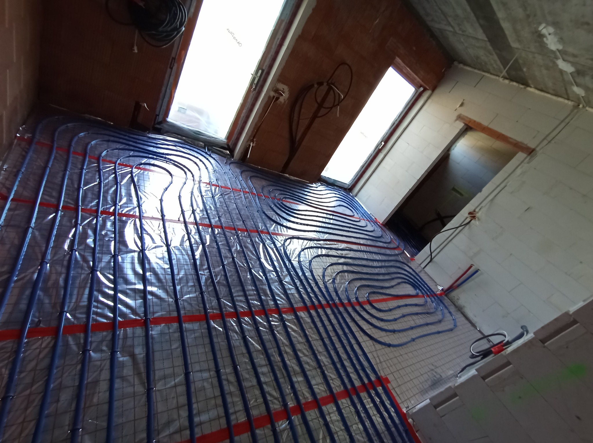 Niebieskie rury ogrzewania podłogowego ułożone na folii izolacyjnej z czerwonymi paskami, widok z góry w surowym wnętrzu z oknami dachowymi.