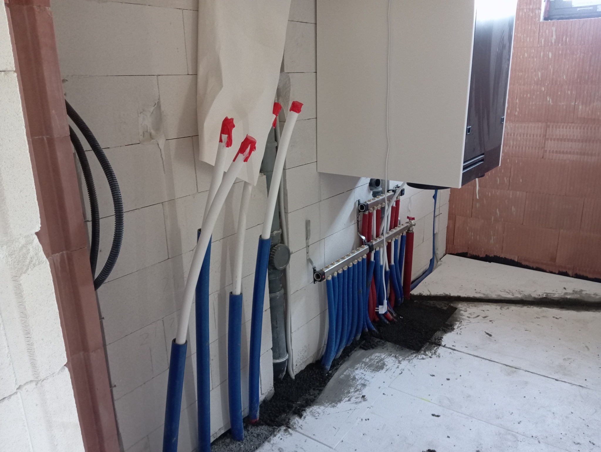Rozdzielacz ogrzewania podłogowego z rurami w izolacji, zamontowany przy ścianie z bloczków, z widocznym piecem gazowym i elementami instalacji.