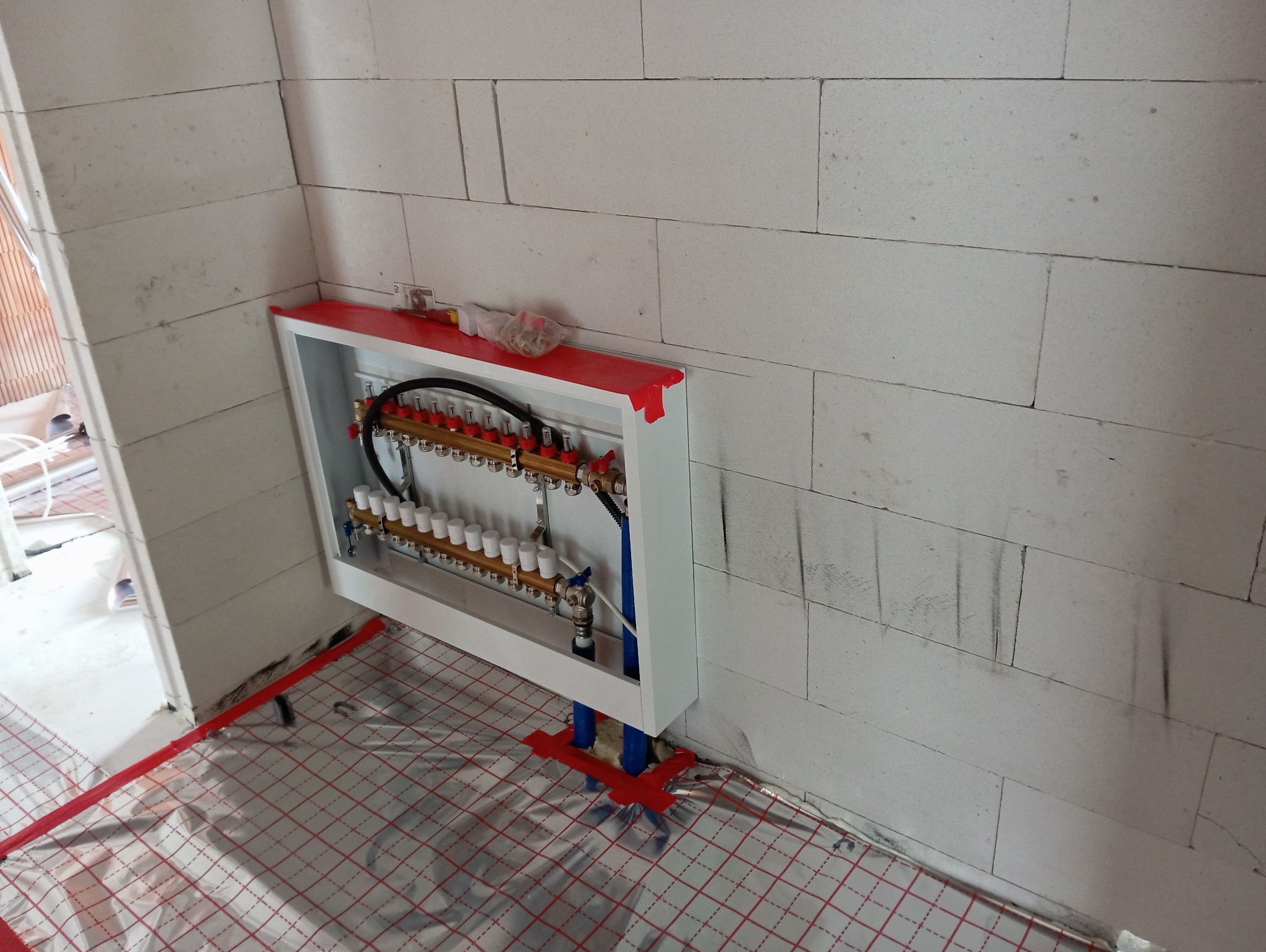 Rozdzielacz ogrzewania podłogowego w obudowie, widoczny na tle ściany z betonu komórkowego i folii izolacyjnej z czerwonym rastrem. Elementy instalacji w kolorze mosiądzu i czerwieni.