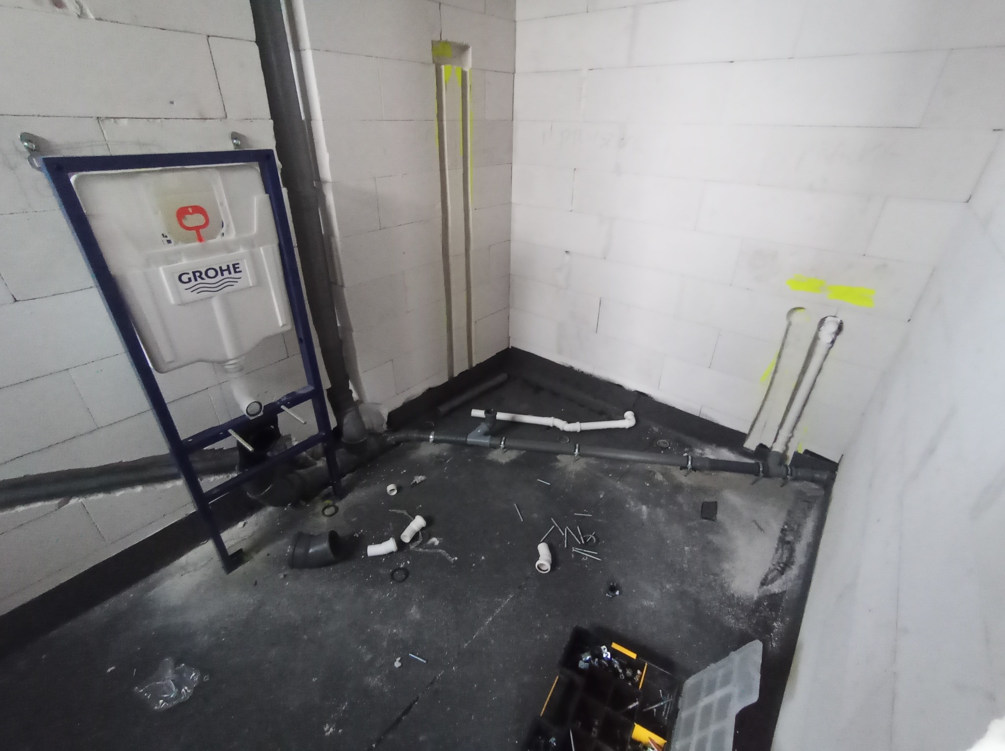 Montaż podtynkowego stelaża WC Grohe w surowej łazience z widocznymi rurami i instalacją kanalizacyjną. W tle ściany z betonu komórkowego.