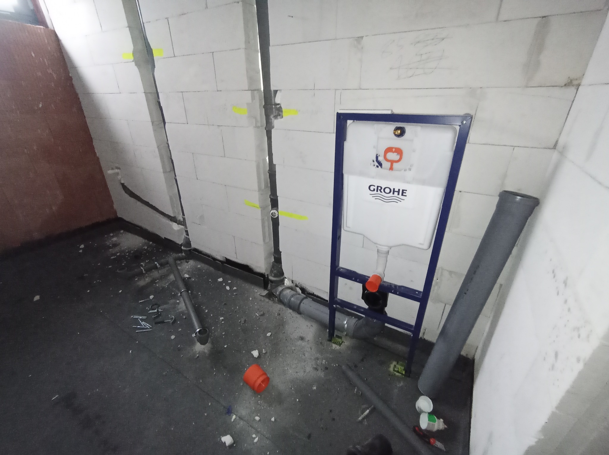 Montaż stelaża podtynkowego Grohe z widocznymi rurami kanalizacyjnymi w surowym wnętrzu z białych bloczków. Żółte oznaczenia na ścianie, podłoga w trakcie prac.