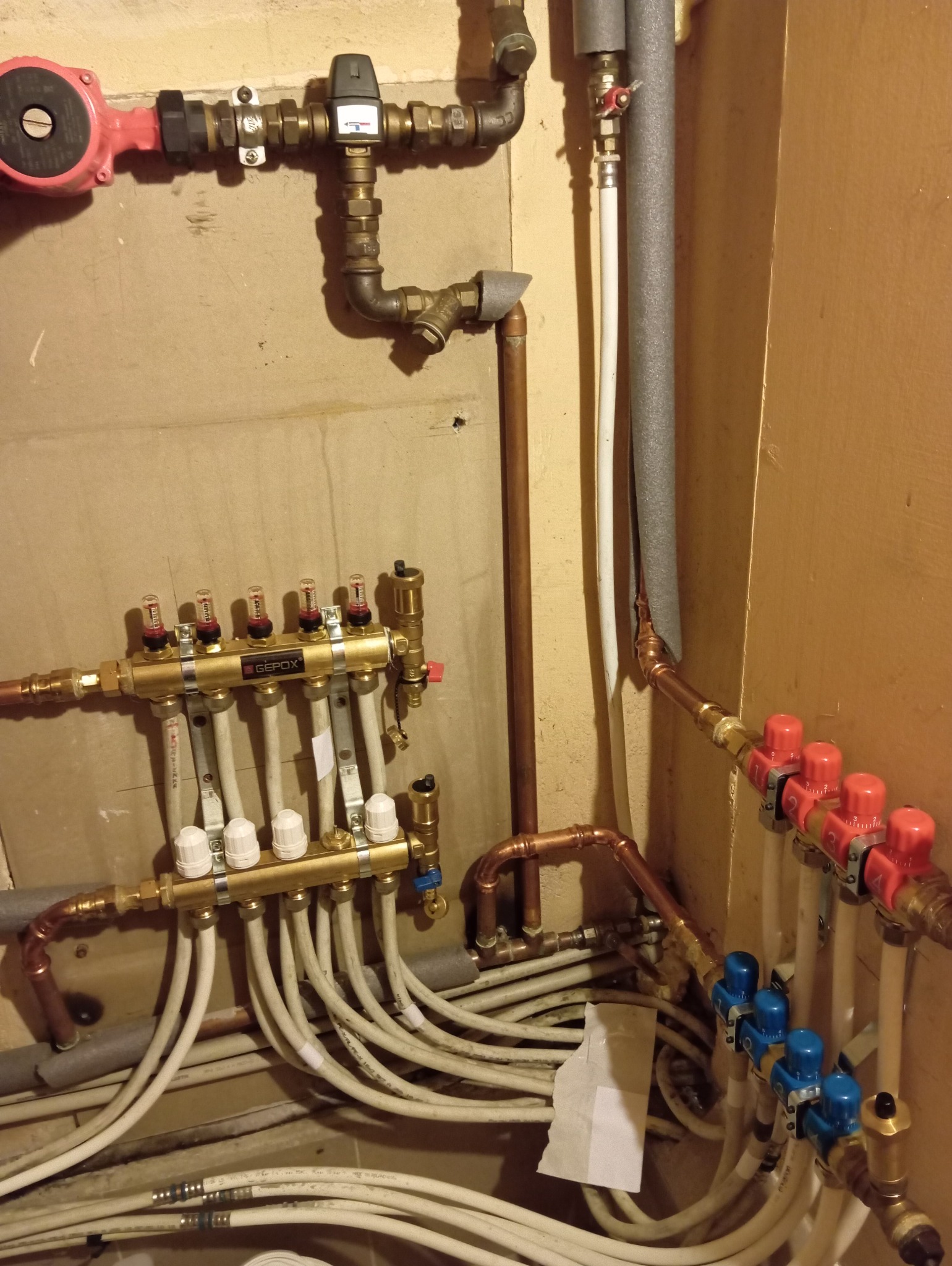 Rozdzielacz ogrzewania podłogowego z rurami, zaworami i elementami instalacji grzewczej na tle ściany. Widoczne złącza i przewody.