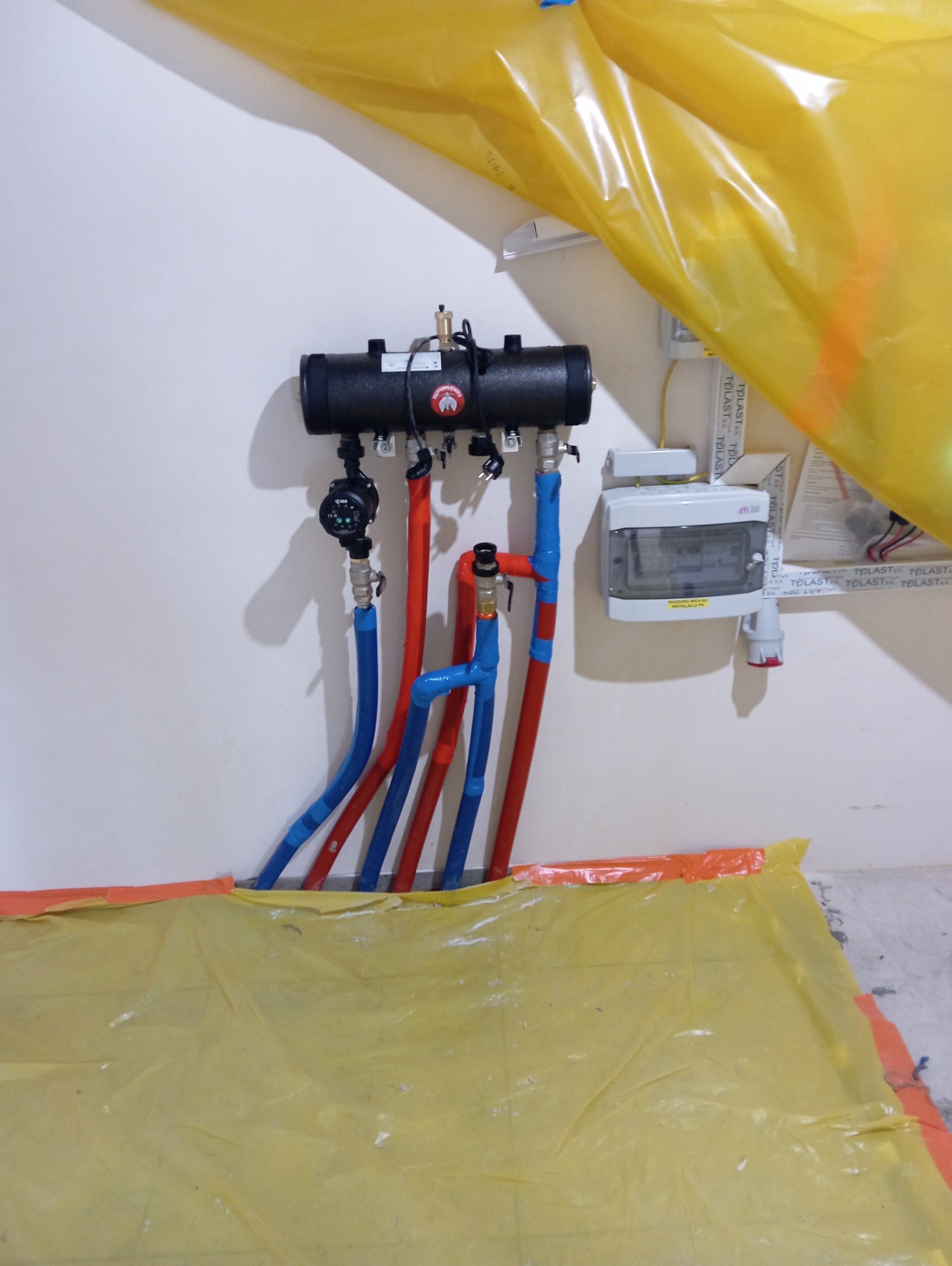 Rozdzielacz ogrzewania podłogowego, z czerwonymi i niebieskimi rurami, zamontowany na białej ścianie, zabezpieczony folią malarską.